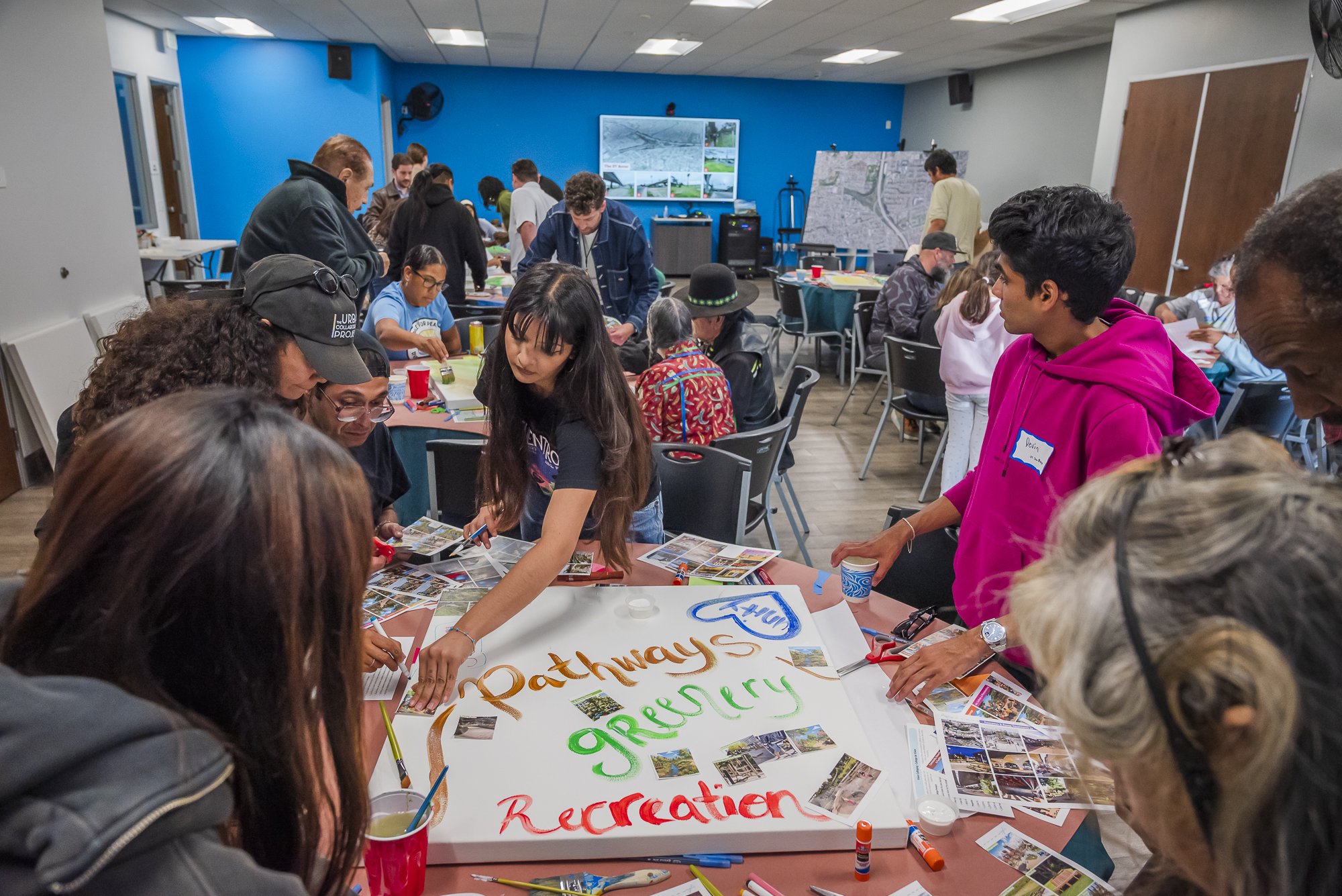 A diverse group of people in a community center room gathered around a large poster on a table. The poster has the words 'Pathways to Recreation' written on it with various photos attached. People are engaging in arts and crafts activities, and some 