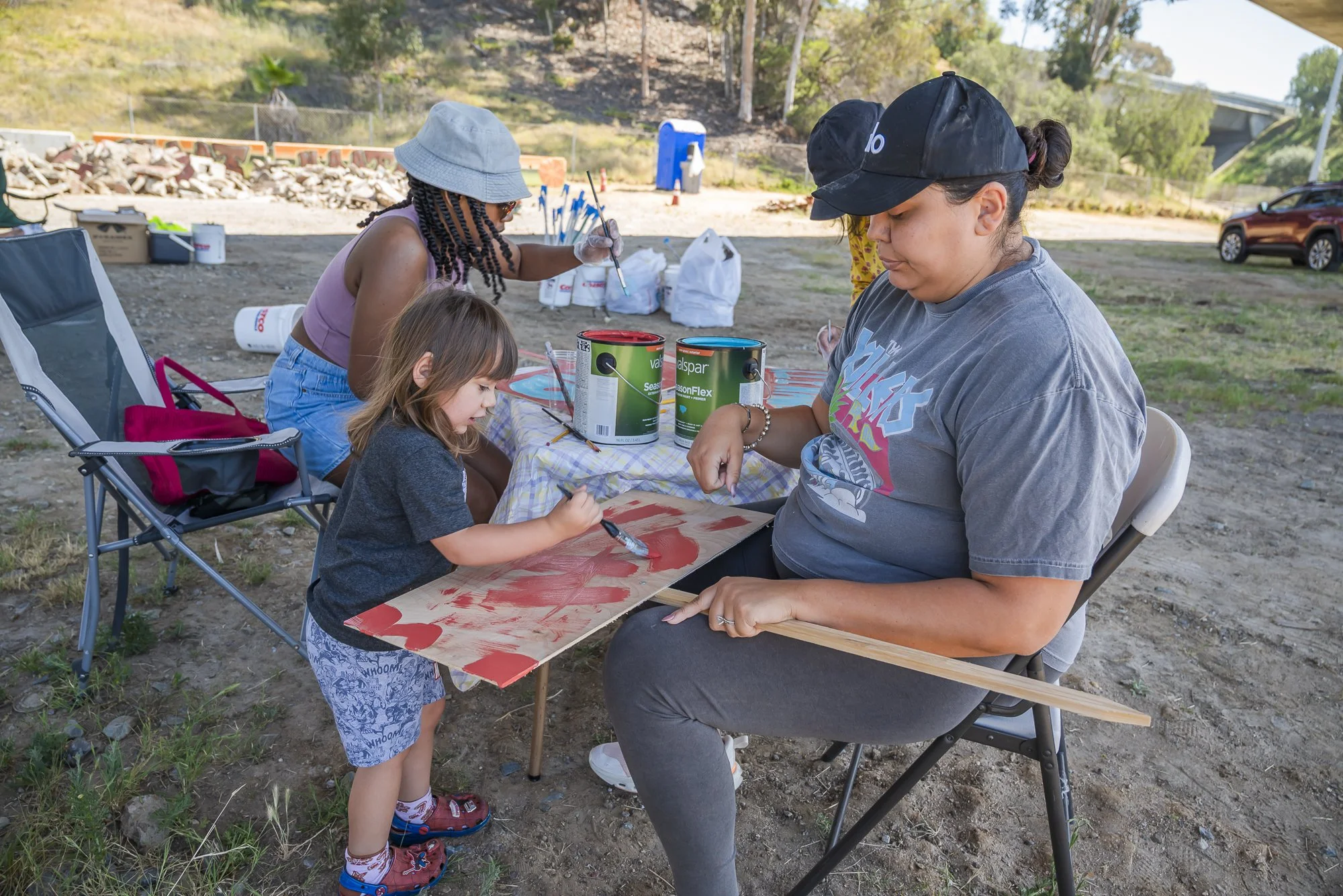 A young girl and a woman painting on a piece of wood at an outdoor event. The girl is focused on her art while the woman, wearing a baseball cap, looks at her work. There are paint cans, brushes, and supplies on the table, with a backdrop of trees, a