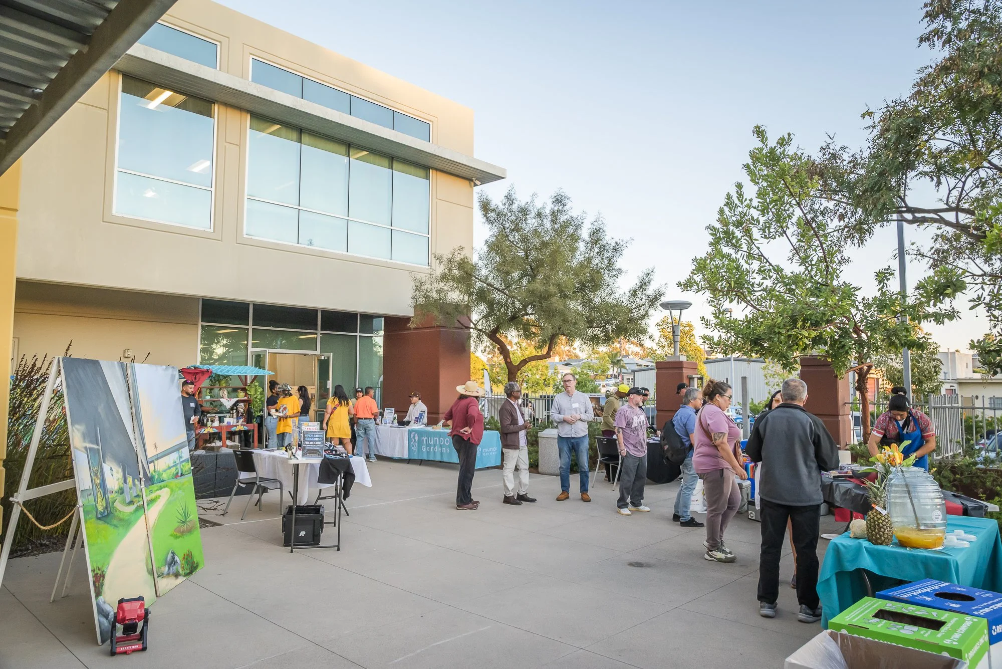 People gather outdoors at a community event with tables and booths set up, some displaying artwork, food, and informational materials, in a courtyard area next to a modern building with large glass windows.