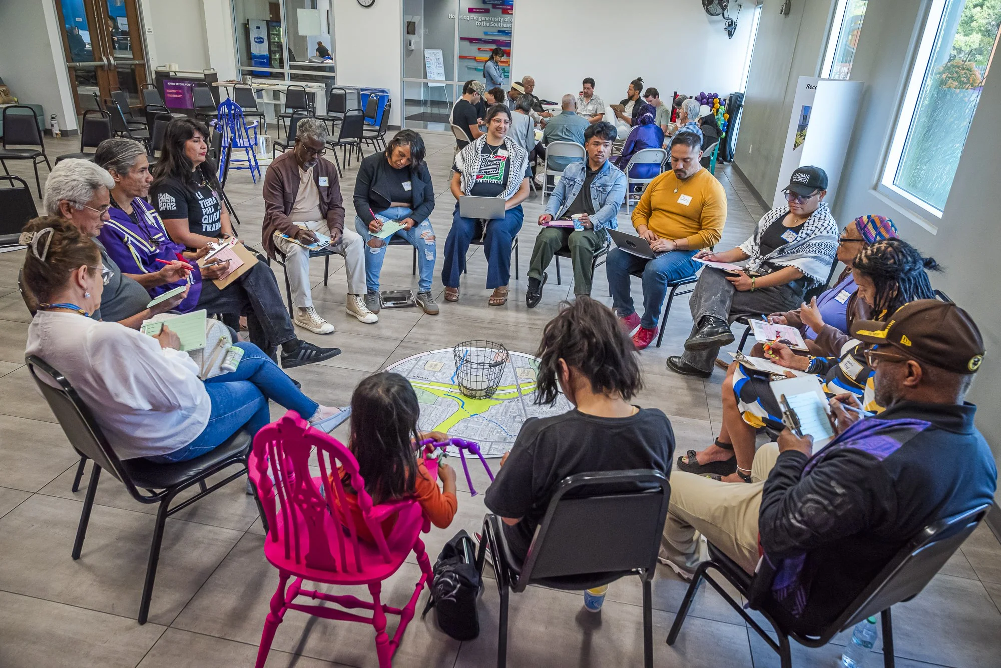 A diverse group of people sitting in a circle at a community event or meeting in a bright indoor space with large windows and a city map on the floor.