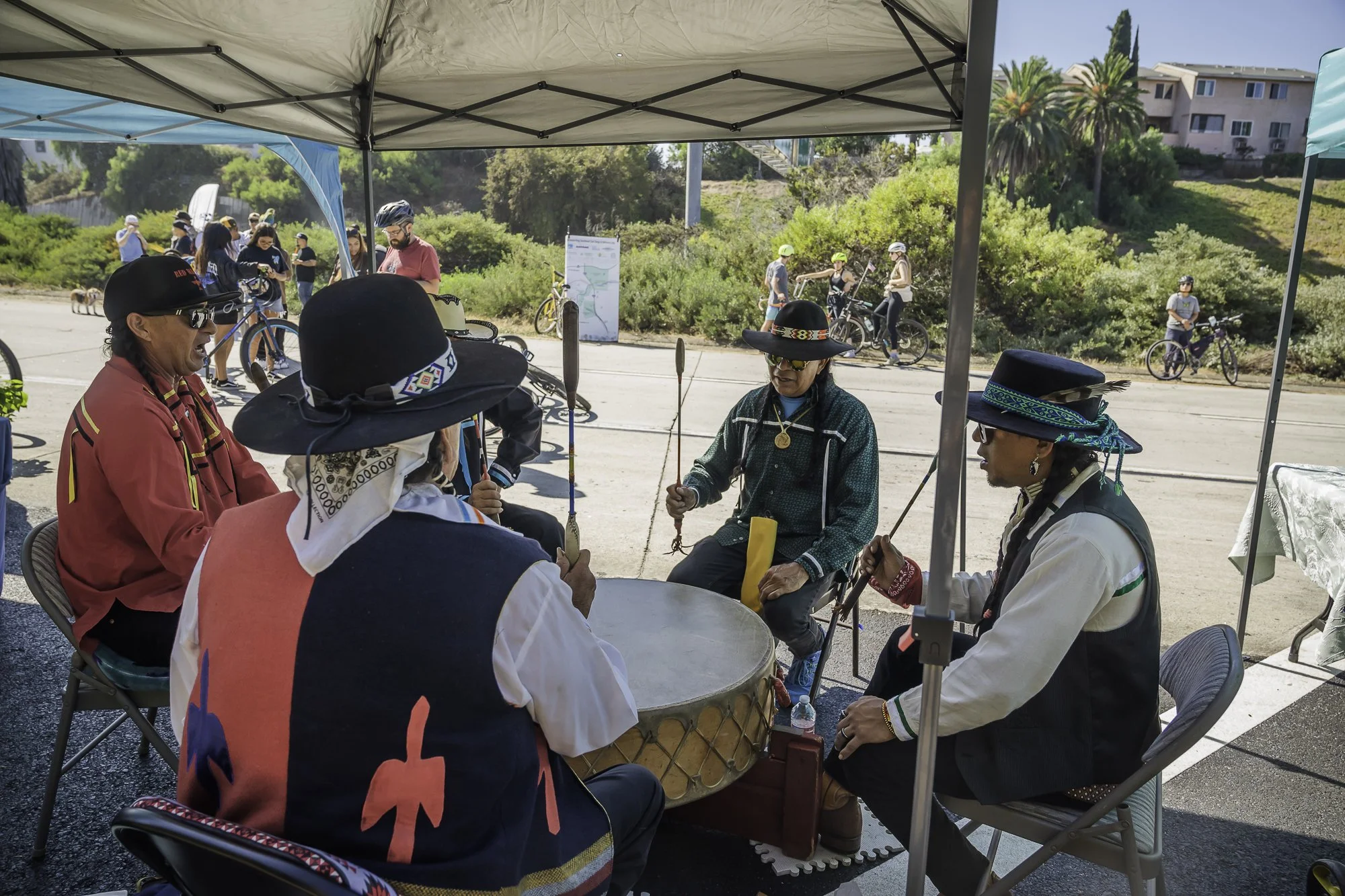 People sitting around a table under a canopy at an outdoor event, with several individuals on bicycles in the background and a hillside with trees and buildings