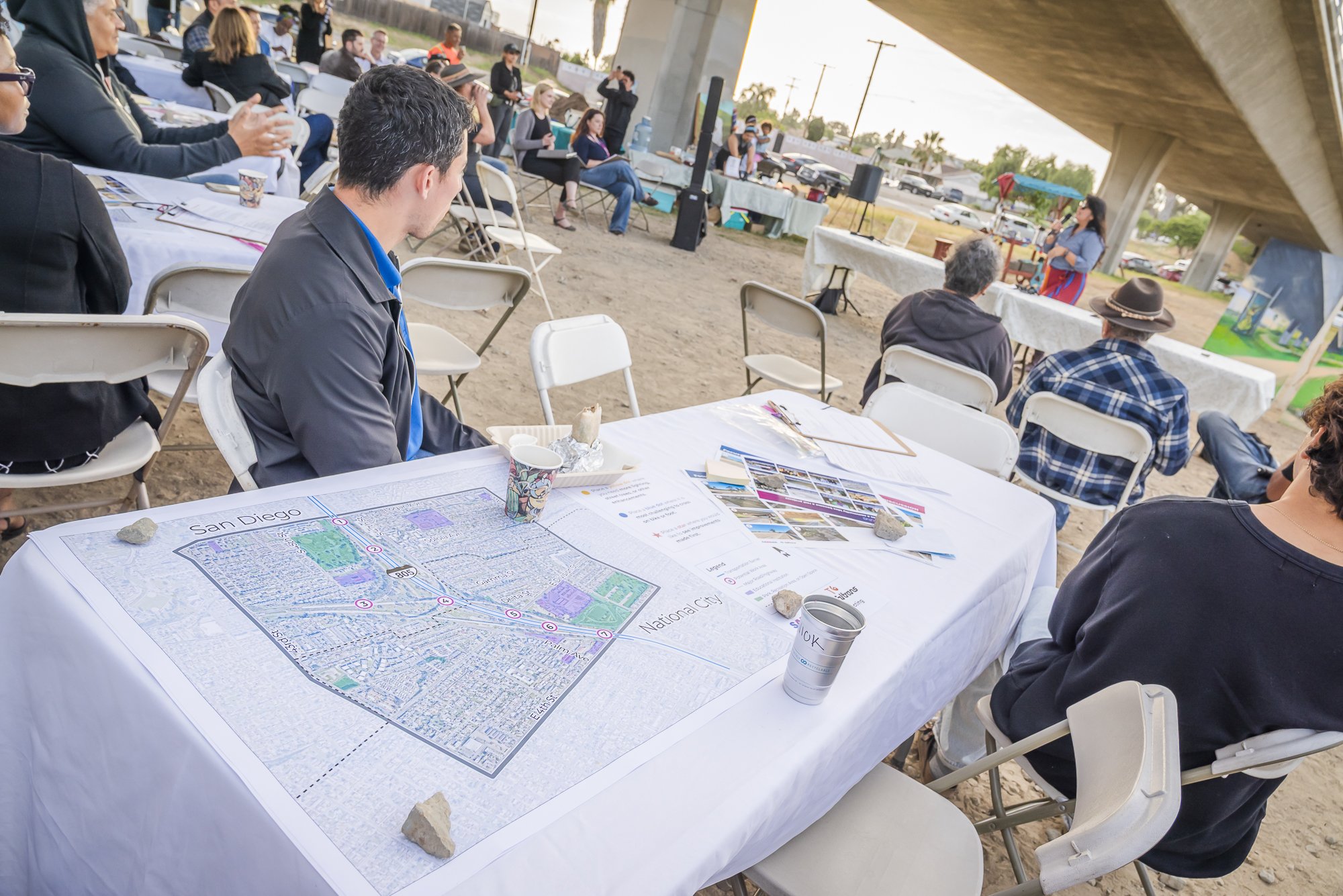 People attending an outdoor presentation or meeting under a bridge, with tables, chairs, and a presenter at the front. A detailed map of San Diego is spread on a table in the foreground.