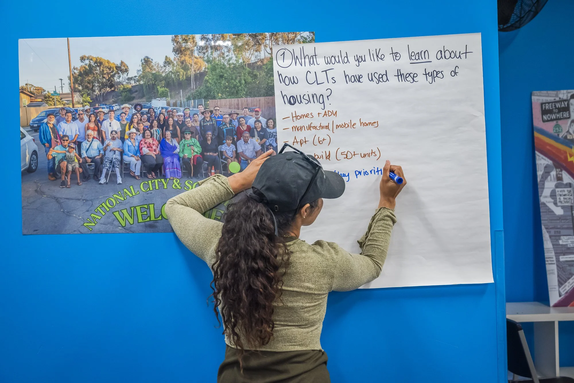 A woman with curly hair, wearing a cap and green jacket, writes on a large white poster. The poster contains a question about how community land trusts have been used, with bullet points listing homes, manufacturing, and apartments. Behind her is a l
