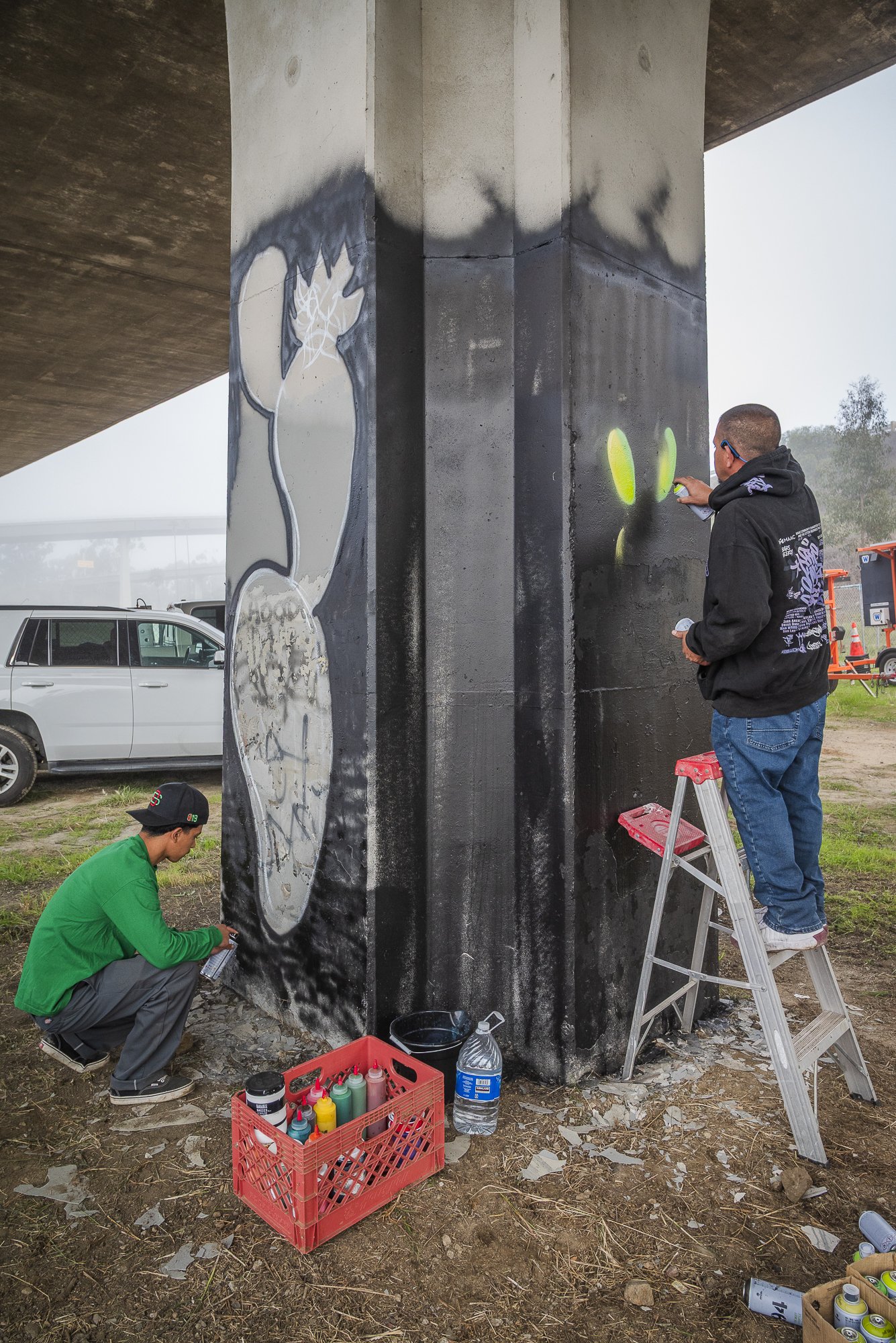 Two artists creating graffiti on a concrete pillar under a bridge. One is standing on a ladder with spray paint, the other is crouching on the ground.