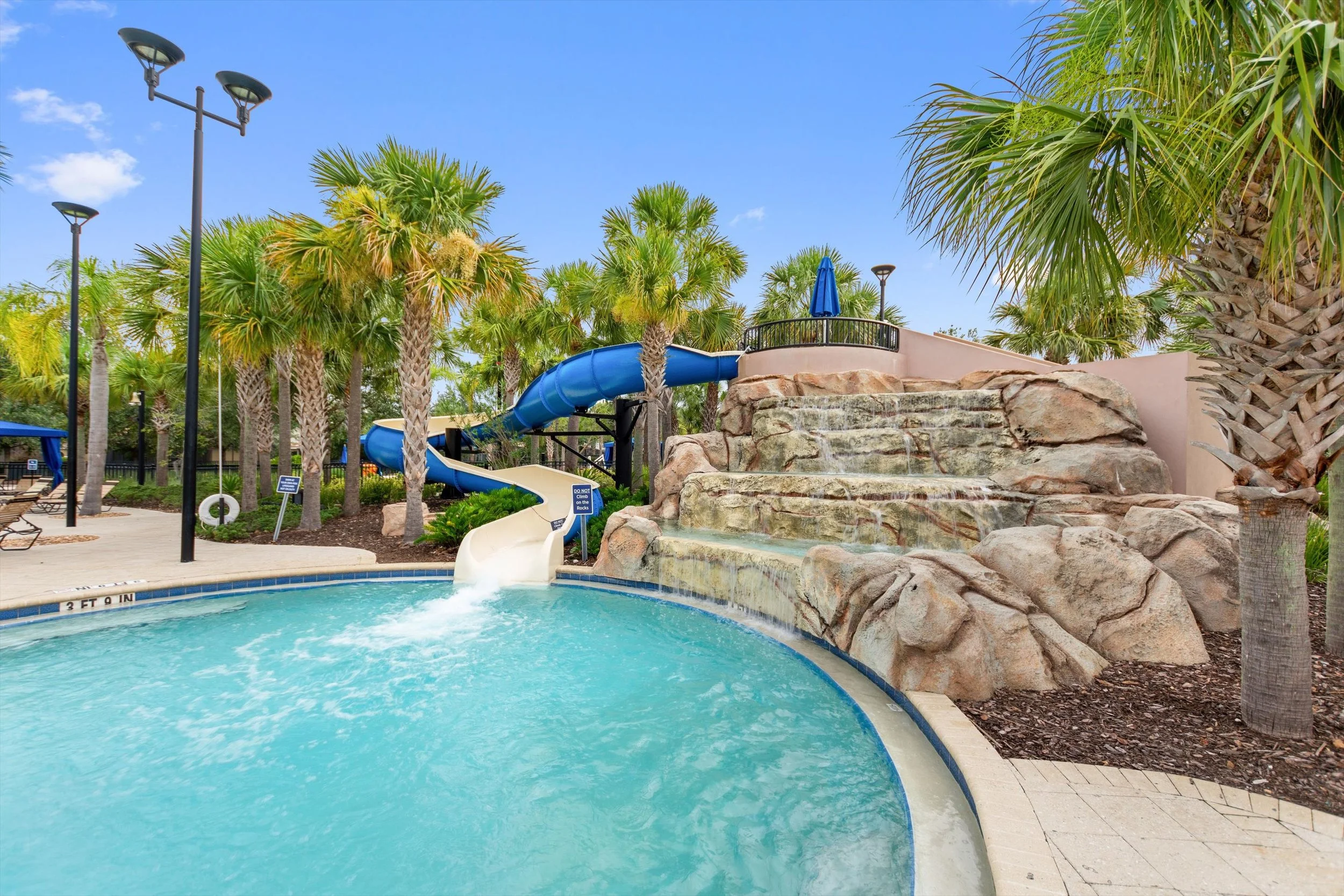 Outdoor swimming pool with a water slide and waterfall feature, surrounded by palm trees and lounge chairs, in a tropical setting under a blue sky.