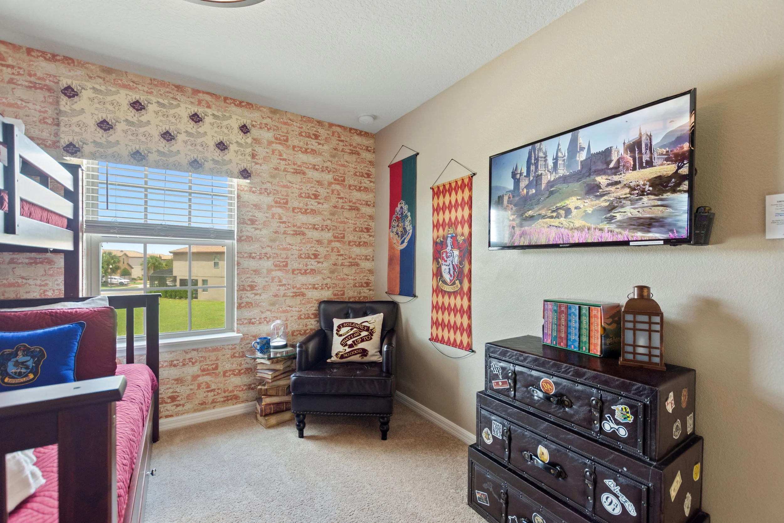 Cozy bedroom with a loft bed, window with blinds, brick accent wall, black armchair, wall decorations including Hogwarts banners, TV showing a castle, and a dresser with stickers and decorative items.
