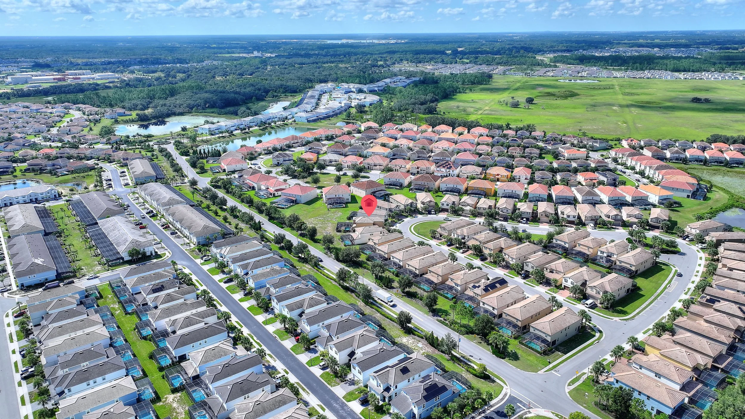 Aerial view of a suburban neighborhood with houses, streets, and green spaces, including a pond and golf course, under a partly cloudy sky.