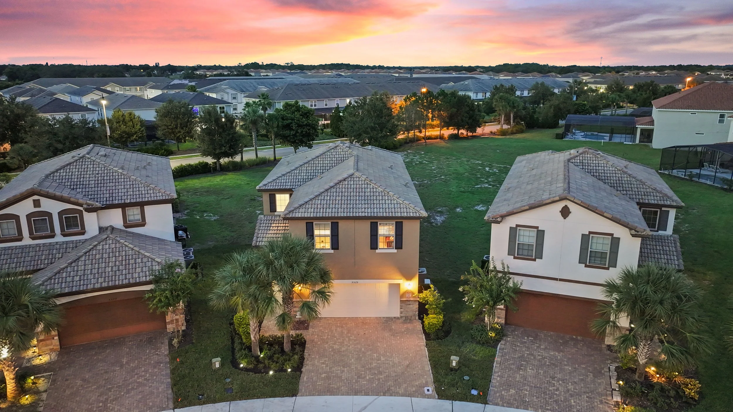 Aerial view of three suburban houses at sunset, with lush green lawns, palm trees, and a back pool area in the background.