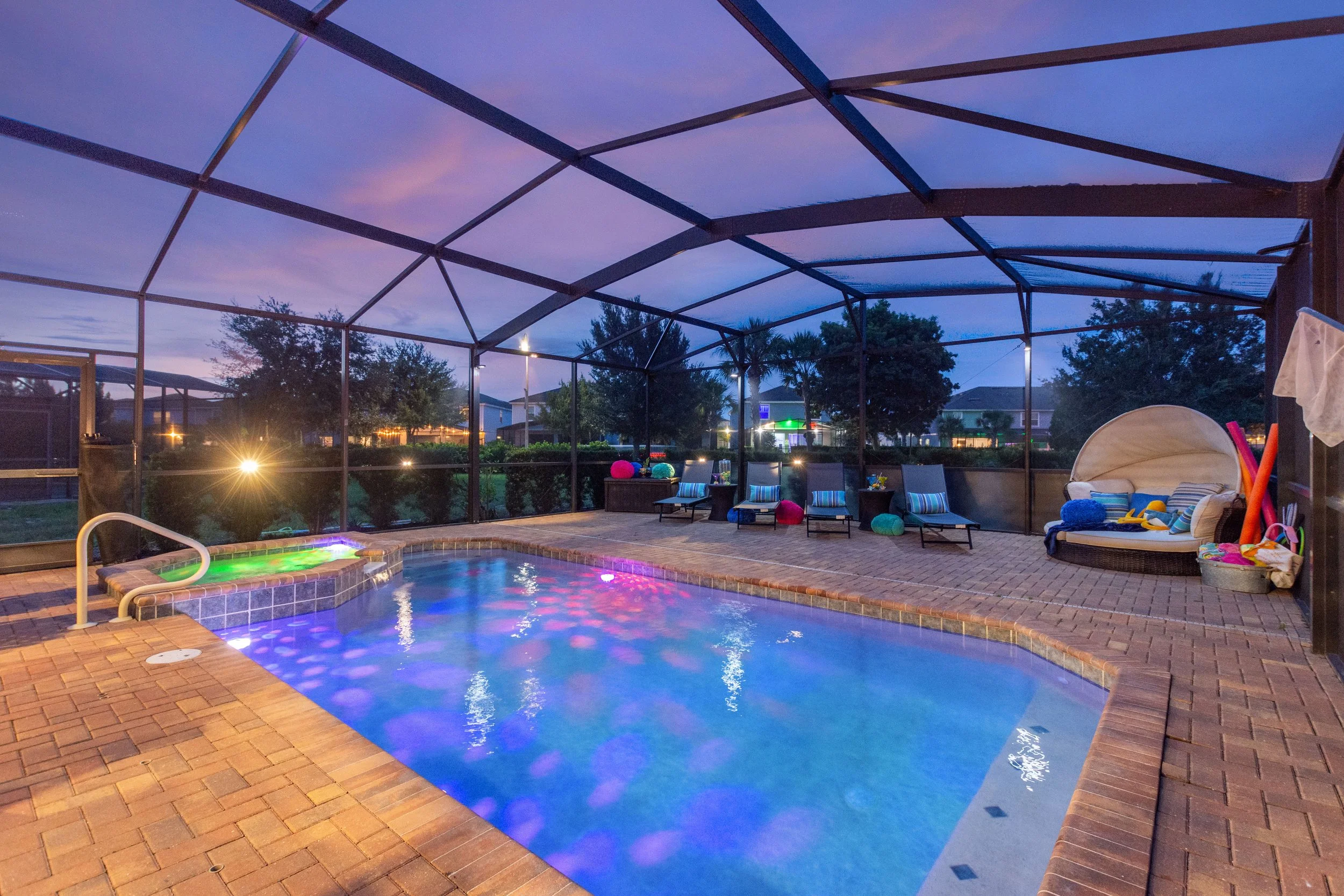 Covered backyard pool area at dusk with chairs, lounge beds, and colorful pool lights.