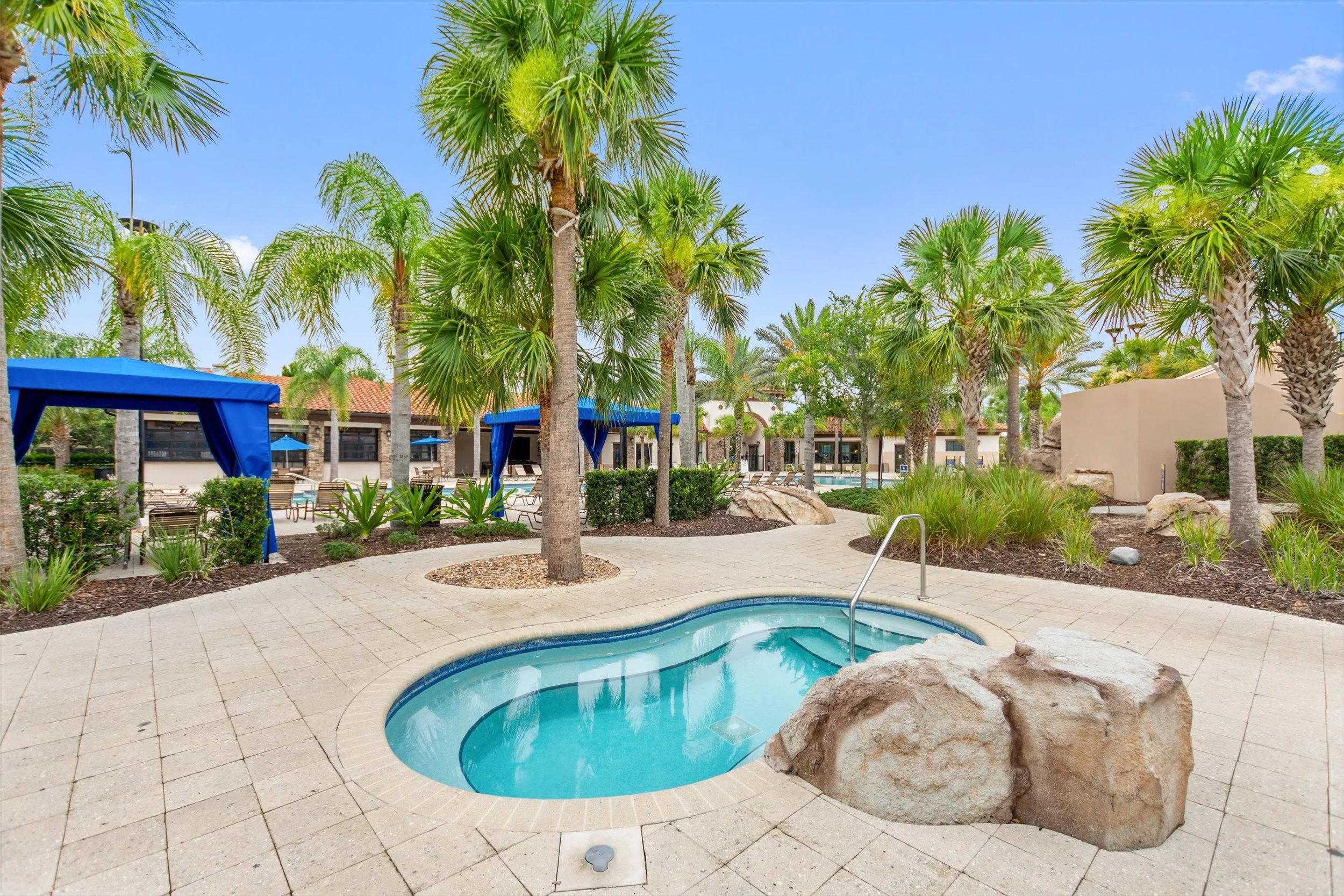 A landscaped poolside area with a small, kidney-shaped pool, surrounded by palm trees, lounge chairs, and shaded cabanas on a bright, sunny day.