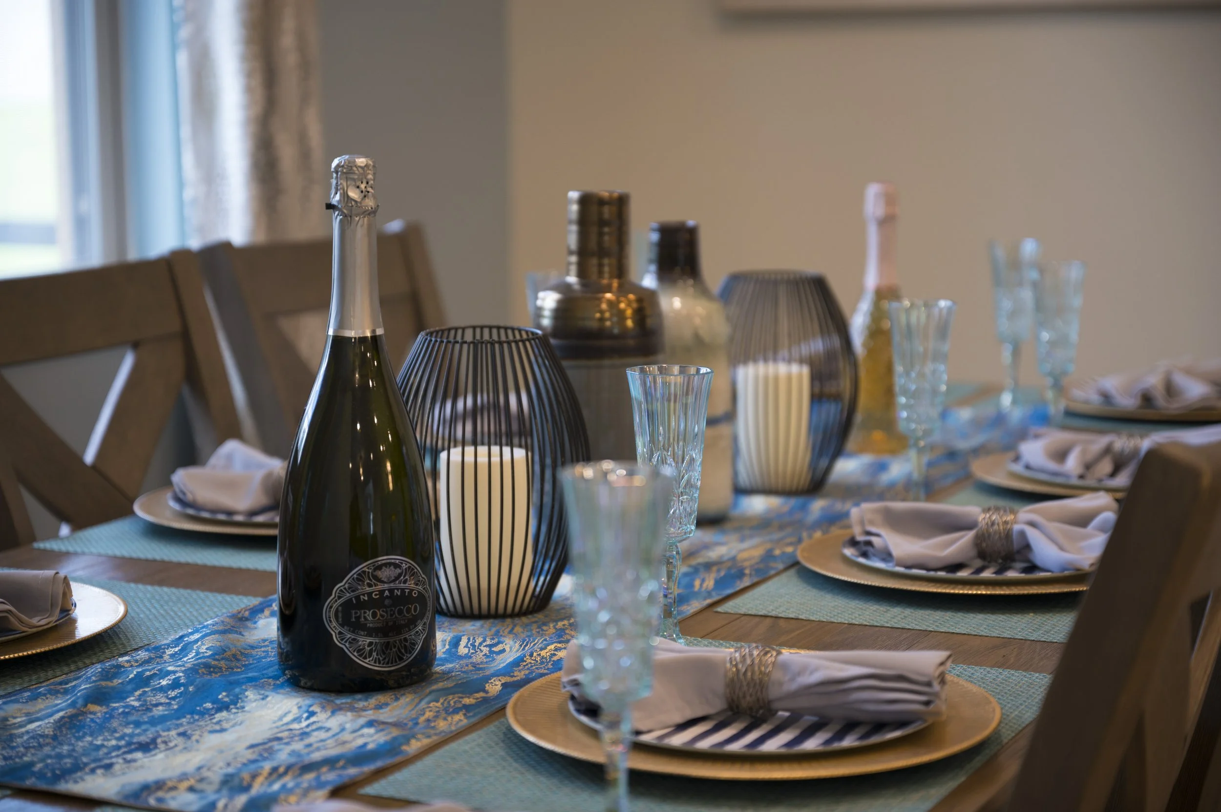 A decorated dining table with a blue and gold table runner, gold chargers, white napkins with napkin rings, tall drinking glasses, candles in black wire lanterns, and bottles of champagne.