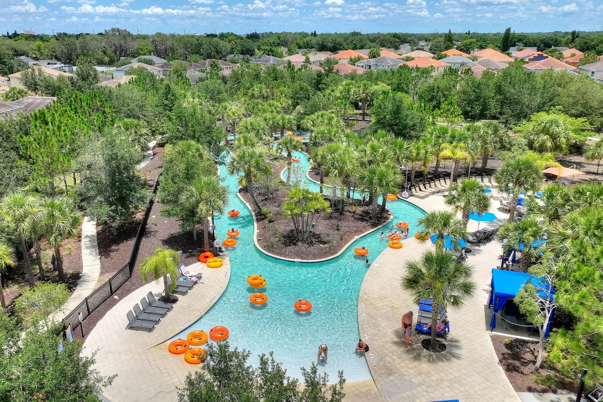 Aerial view of a resort swimming pool area surrounded by lush green trees and residential houses with orange-tiled roofs in the background. The pool has a winding shape with orange float rings and people swimming. There are sun loungers and umbrellas