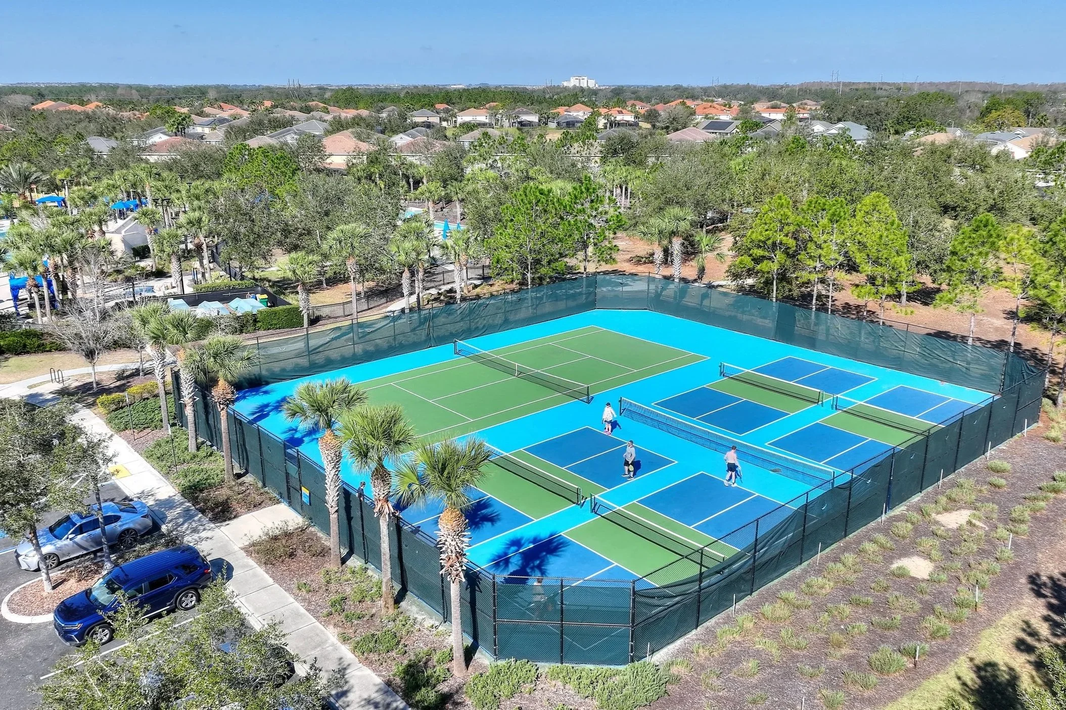 Aerial view of a multi-court tennis and pickleball complex surrounded by trees, with three people playing pickleball on one of the courts, located in a residential community