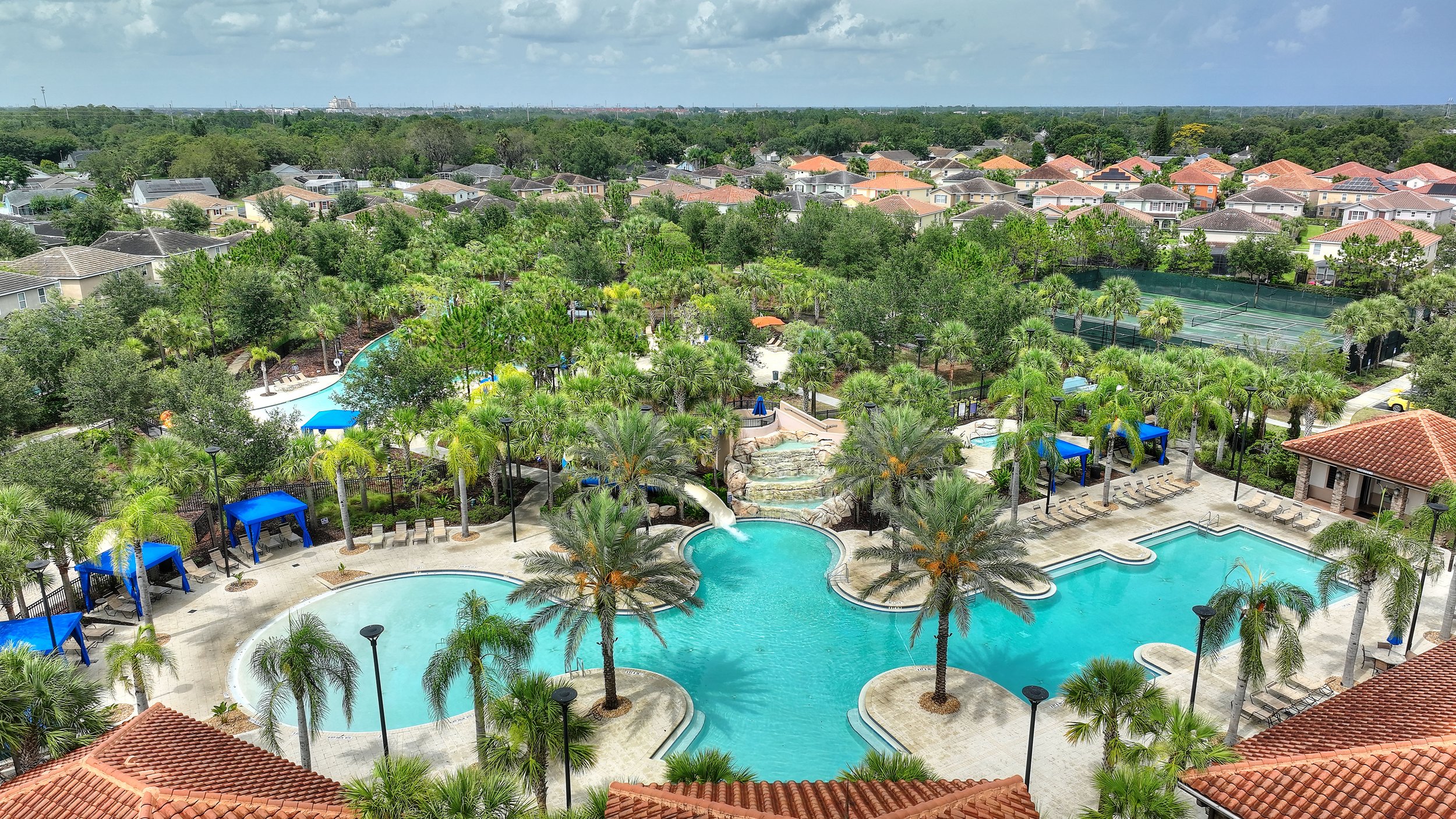 Aerial view of a resort-style swimming pool area with palm trees, lounge chairs, shaded cabanas, and tennis courts surrounded by residential houses and lush greenery under a partly cloudy sky.