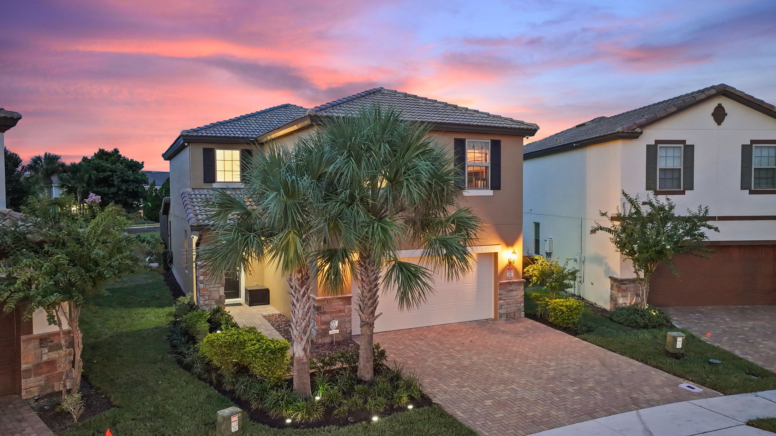 A modern two-story house with a brick driveway, tall palm trees in front, and warm lights on at dusk, with a colorful sunset sky in background.