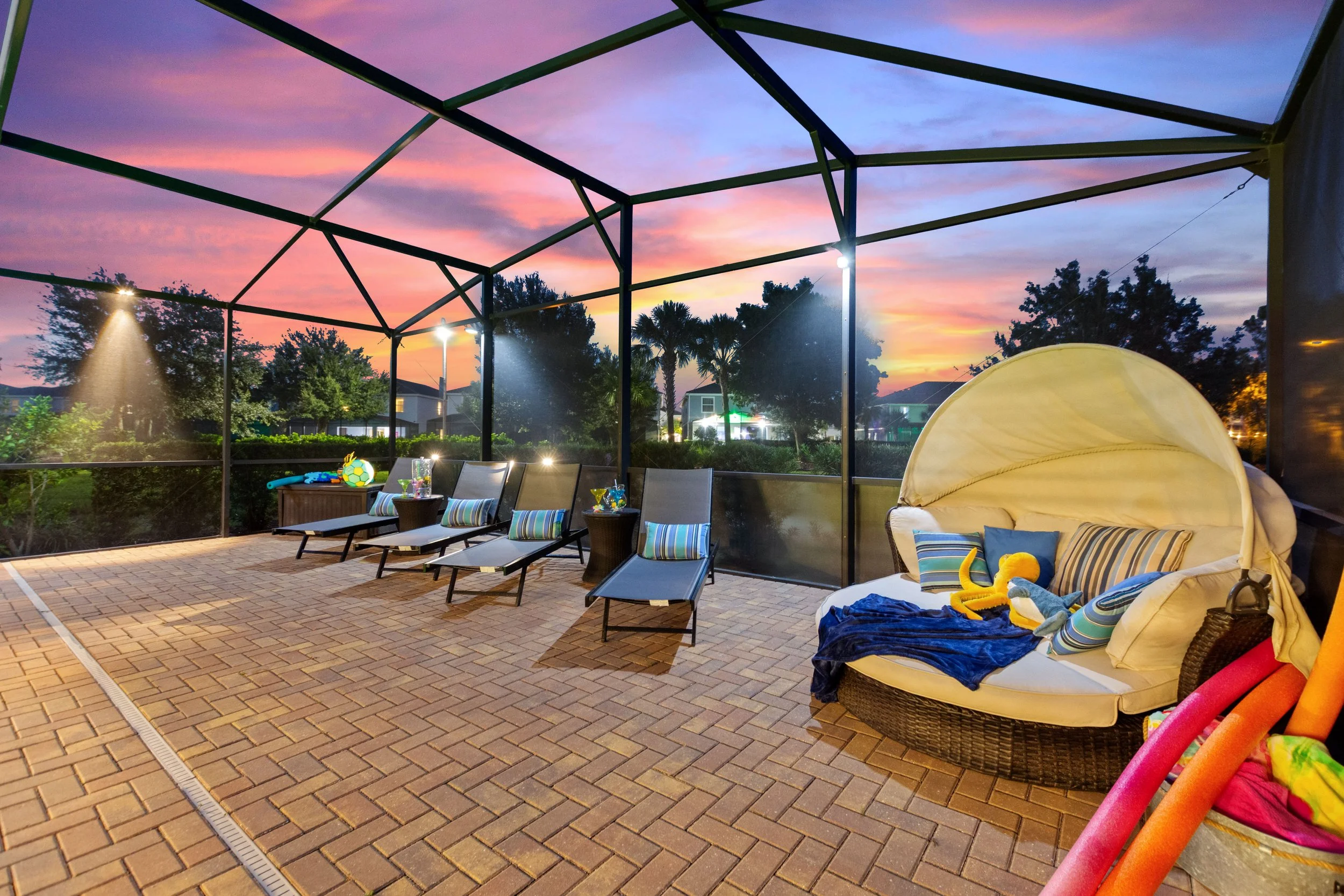Enclosed outdoor patio with lounge chairs and a daybed under a sunset sky, decorated for a poolside setting with pool toys and colorful cushions.