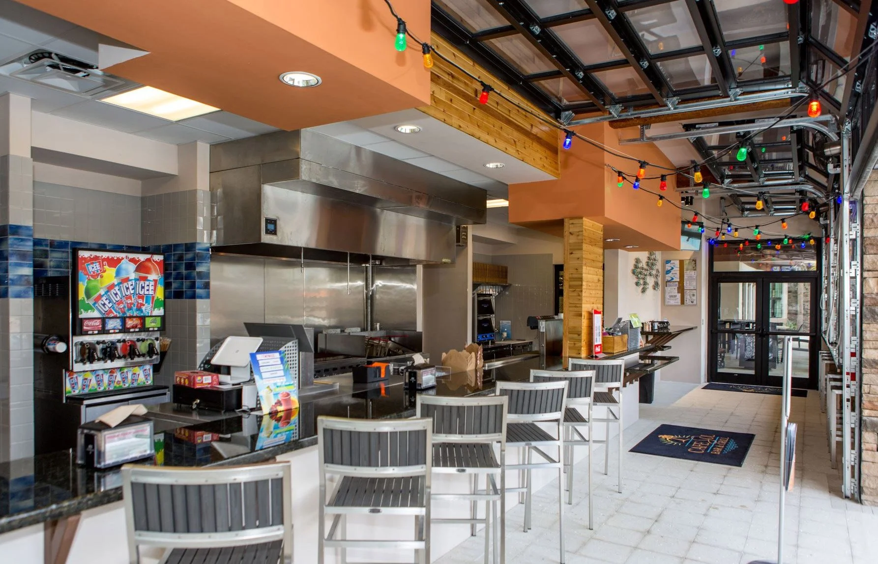 Interior of a casual restaurant or cafe with a counter, barstools, colorful string lights, and a door leading outside.