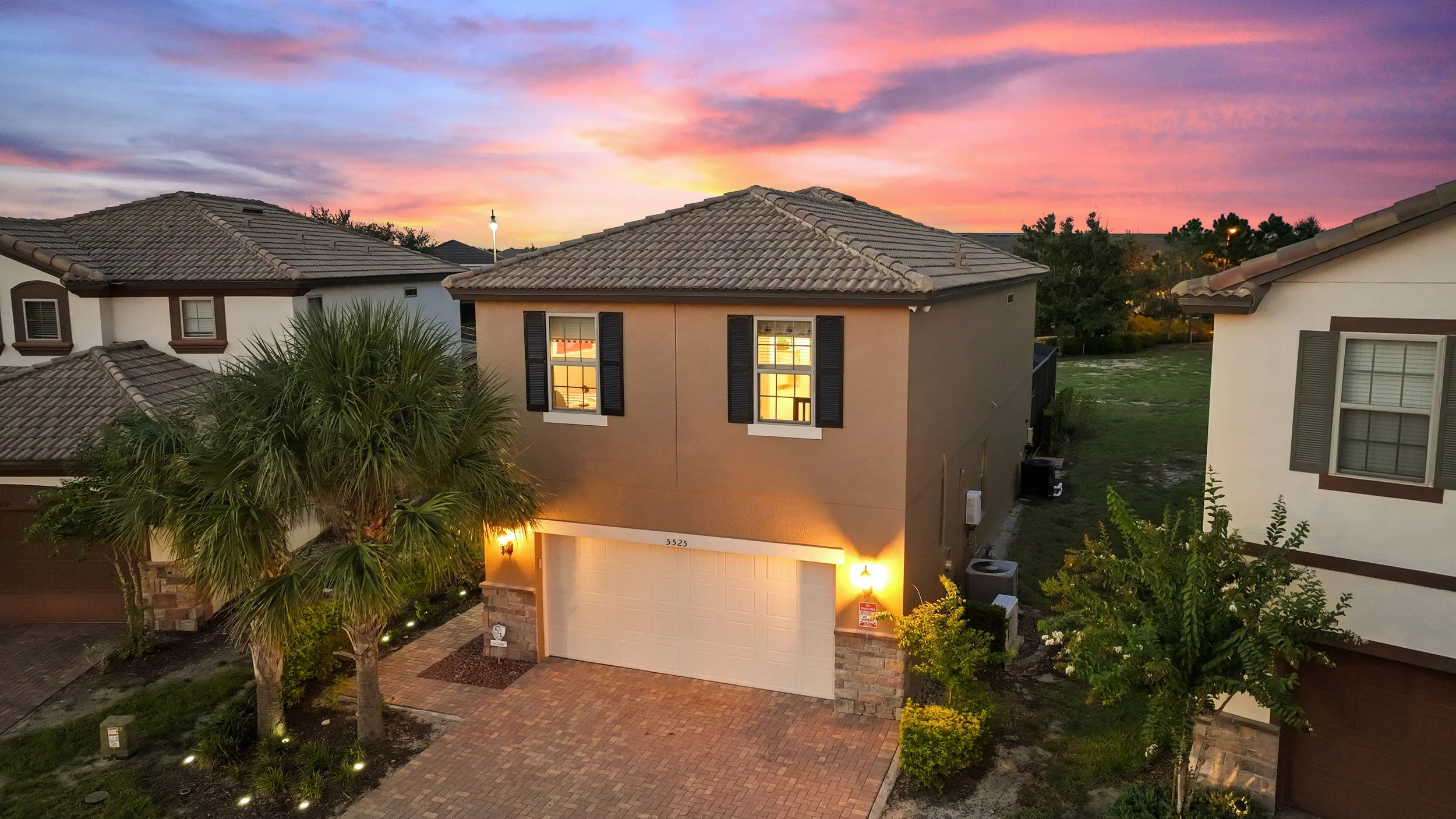 A two-story suburban house during sunset with a brick driveway, palm trees, and lit exterior lights.