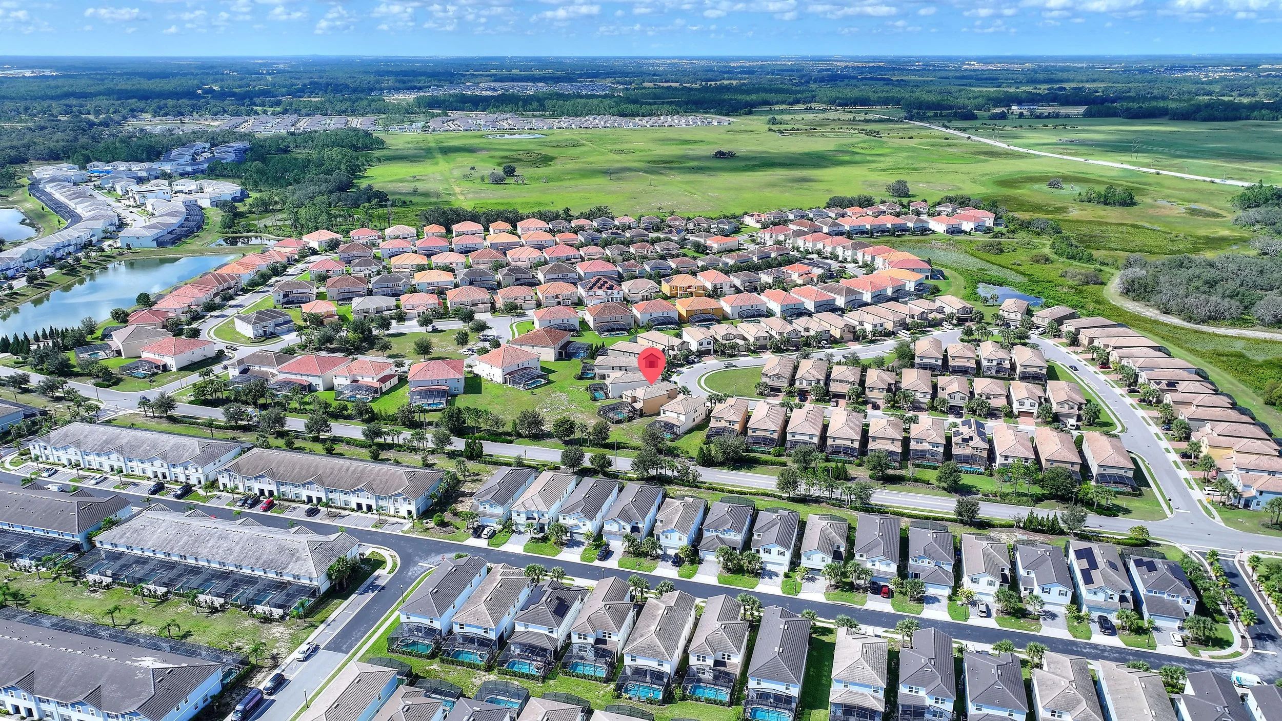 Aerial view of a suburban neighborhood with rows of houses, some with pools, surrounded by greenery and open fields, under a clear blue sky.