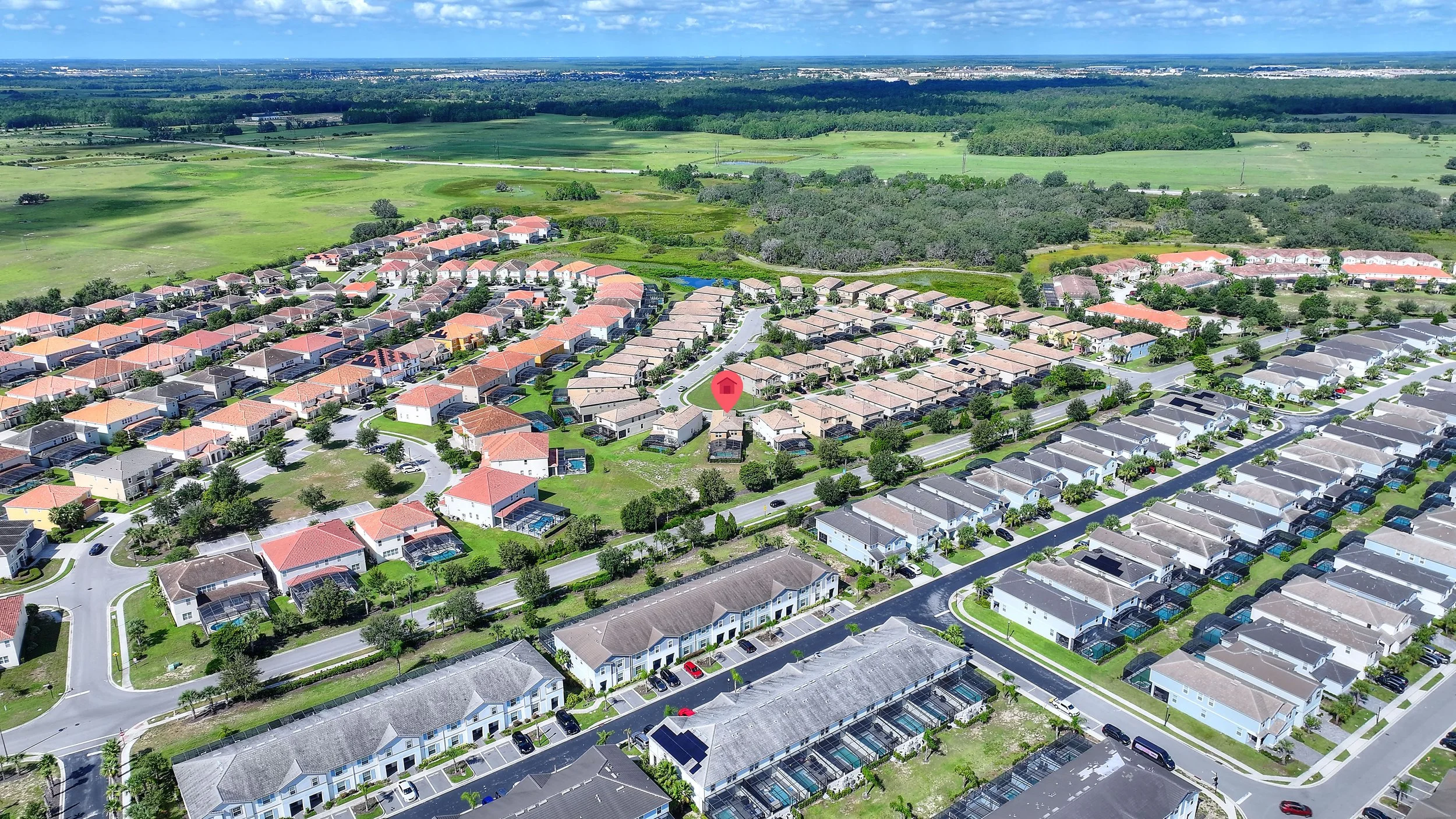 Aerial view of a suburban neighborhood with rows of houses, green lawns, and streets, surrounded by open green fields and distant trees under a partly cloudy sky.