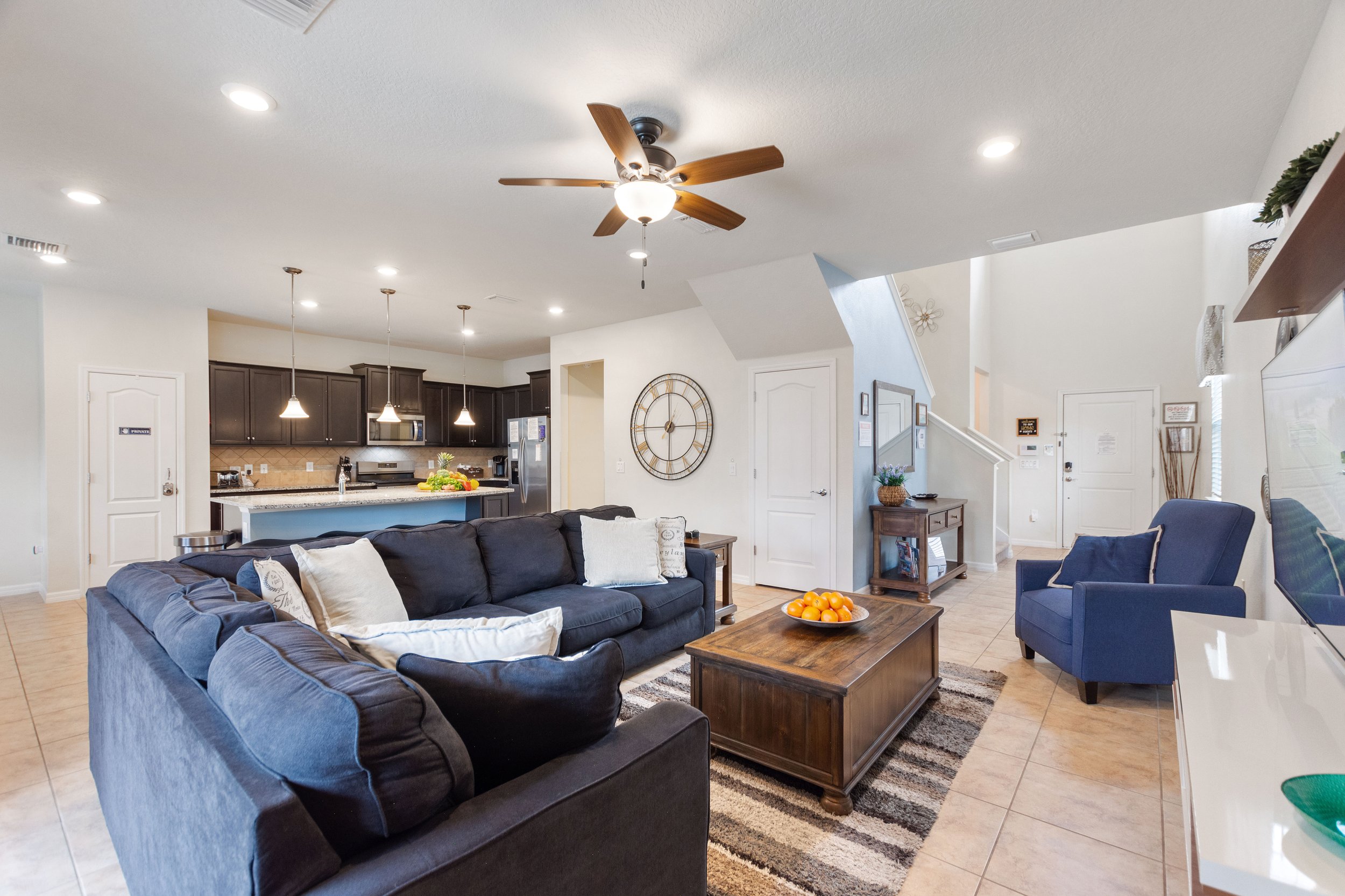 Living room with dark blue sofas, a wooden coffee table with a bowl of oranges, a blue armchair, and a television. The background features an open kitchen with dark cabinets, a kitchen island, and hanging pendant lights. The space has tiled floors, a