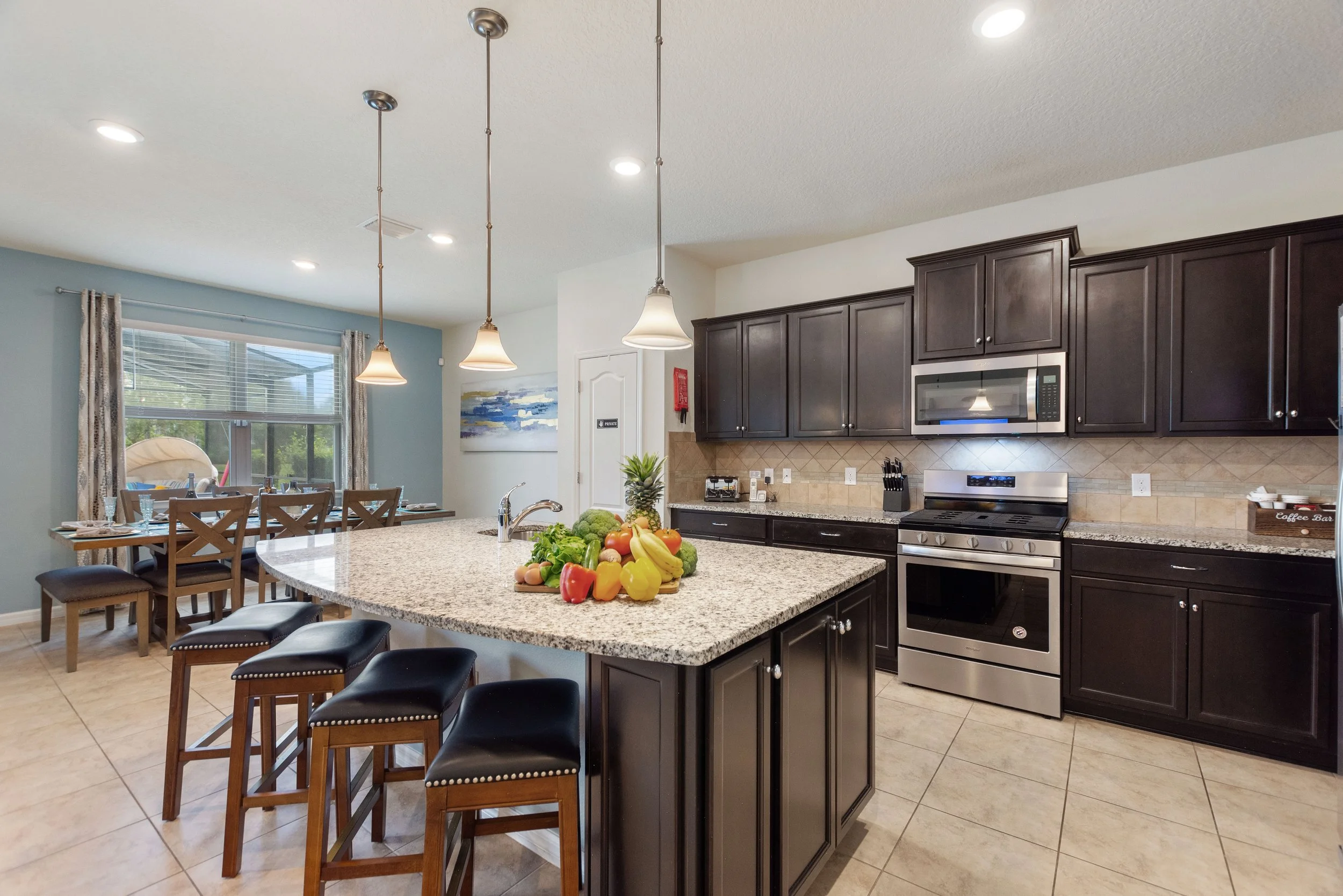 Modern kitchen with dark cabinets, granite countertops, and stainless steel appliances. A kitchen island with a sink and fruits on top, and wooden barstools. Open dining area with a table and chairs near a window.