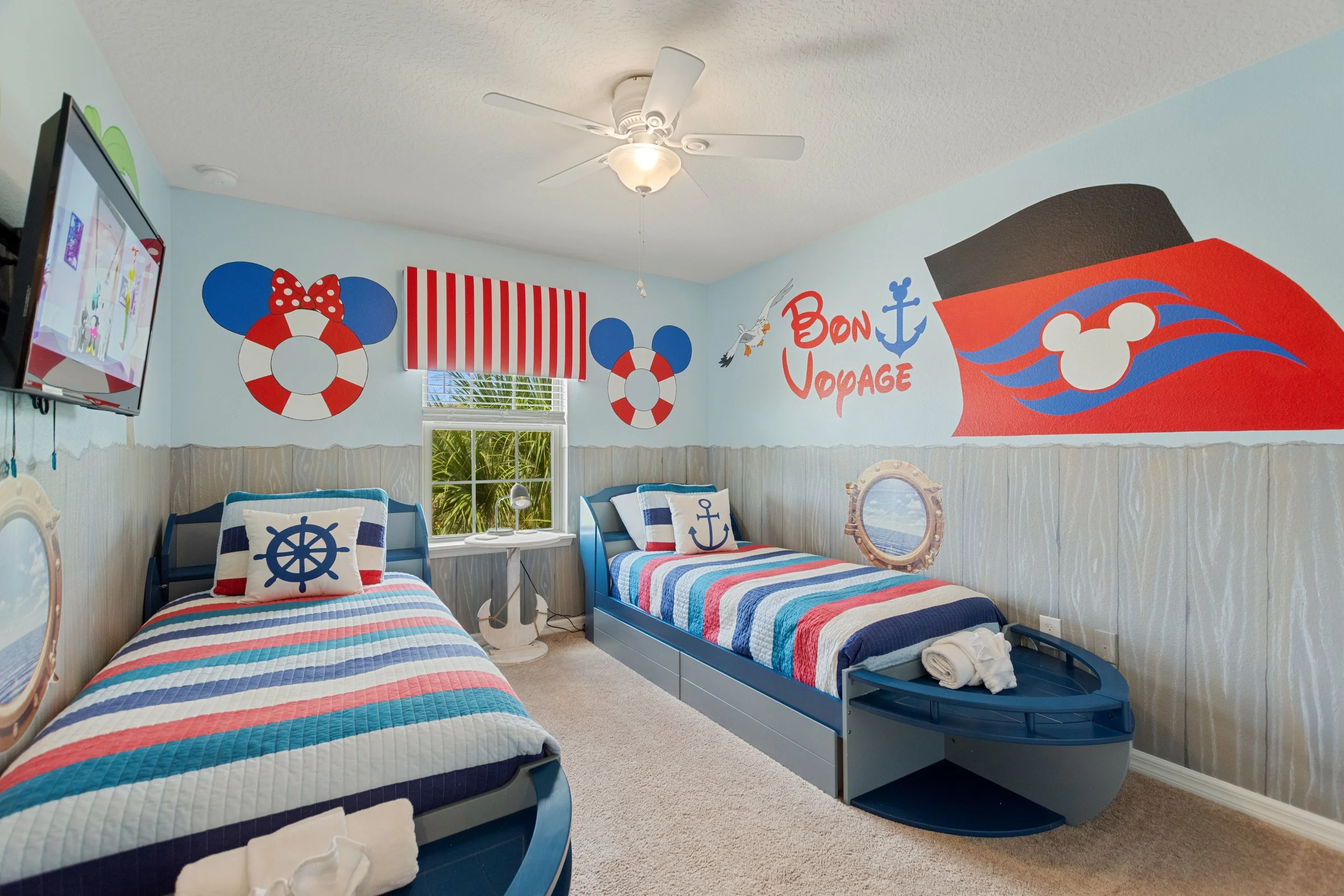 Beach-themed children's bedroom with two twin beds, nautical decor, wall art with a ship and Mickey Mouse helmet, striped comforters, anchor pillows, and a television on the wall.