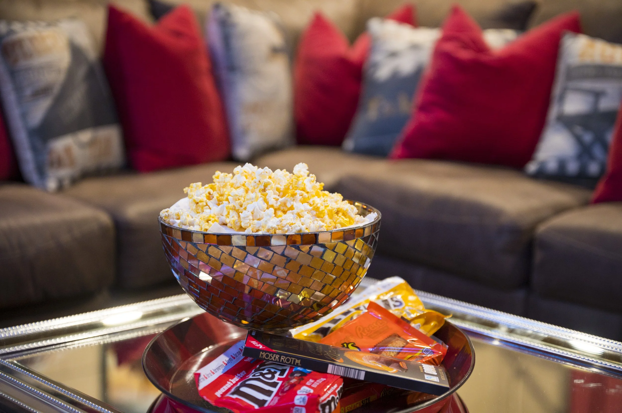A mosaic-patterned bowl filled with popcorn on a silver tray, with snack packages underneath, on a mirrored table in a living room with a sofa and red and patterned pillows in the background.