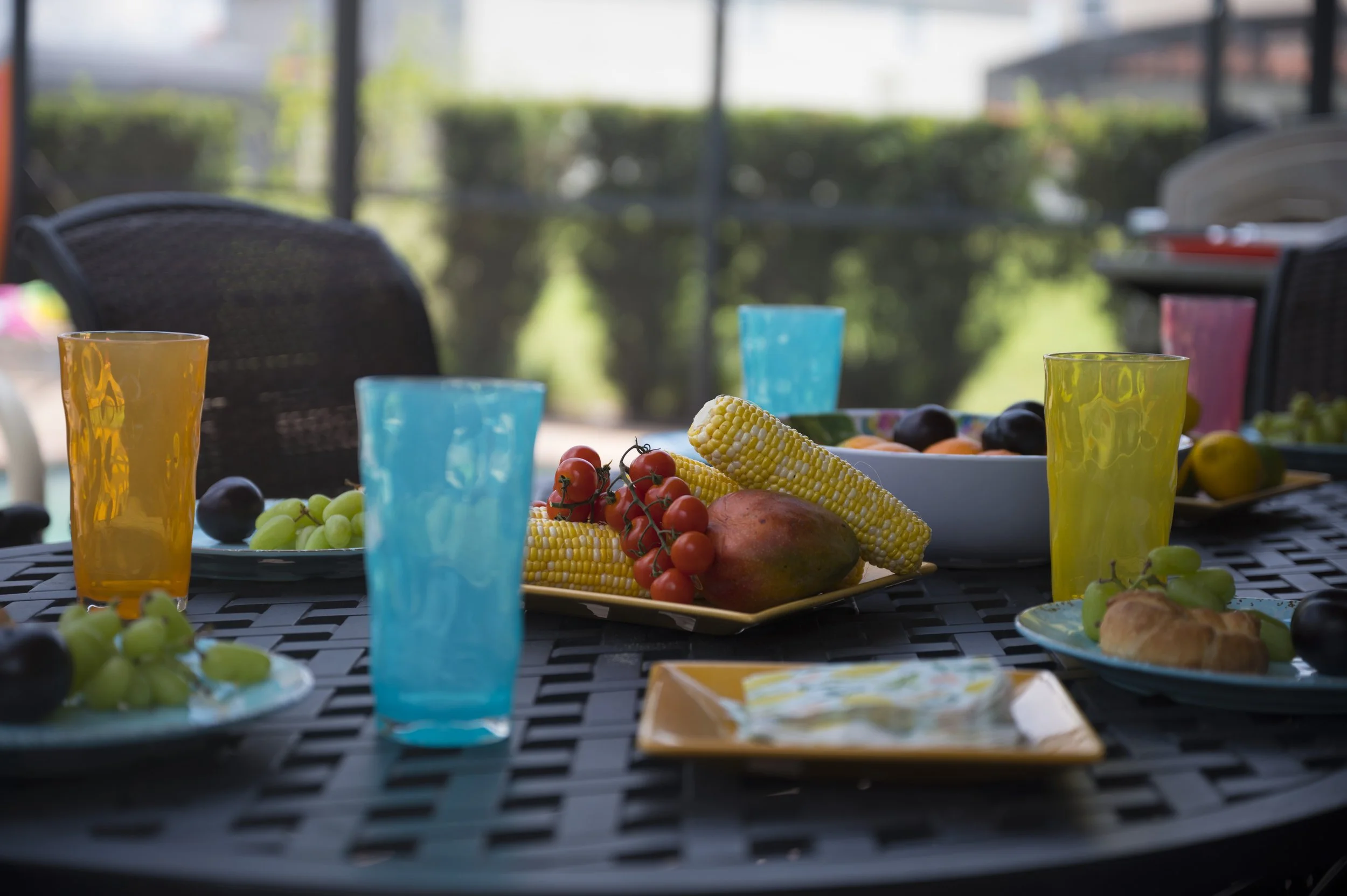 A table set for an outdoor meal with colorful drink glasses, plates of fresh fruit including grapes, cherry tomatoes, corn on the cob, and a mango, as well as a croissant, on a black metal table with a patio and green hedge in the background.