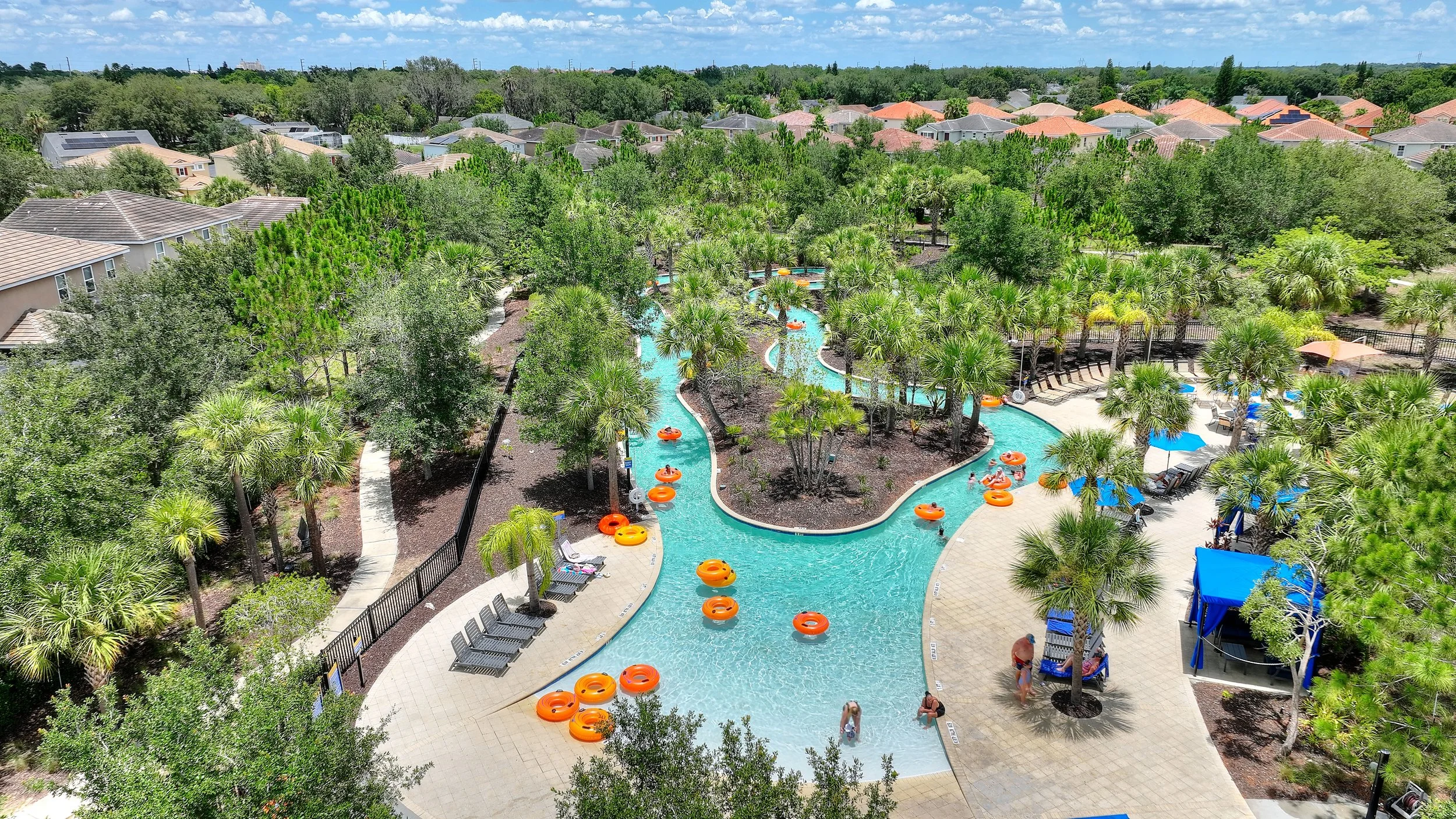Resort swimming pool with winding water slide, surrounded by palm trees, lounge chairs, umbrellas, and houses in the background on a sunny day.