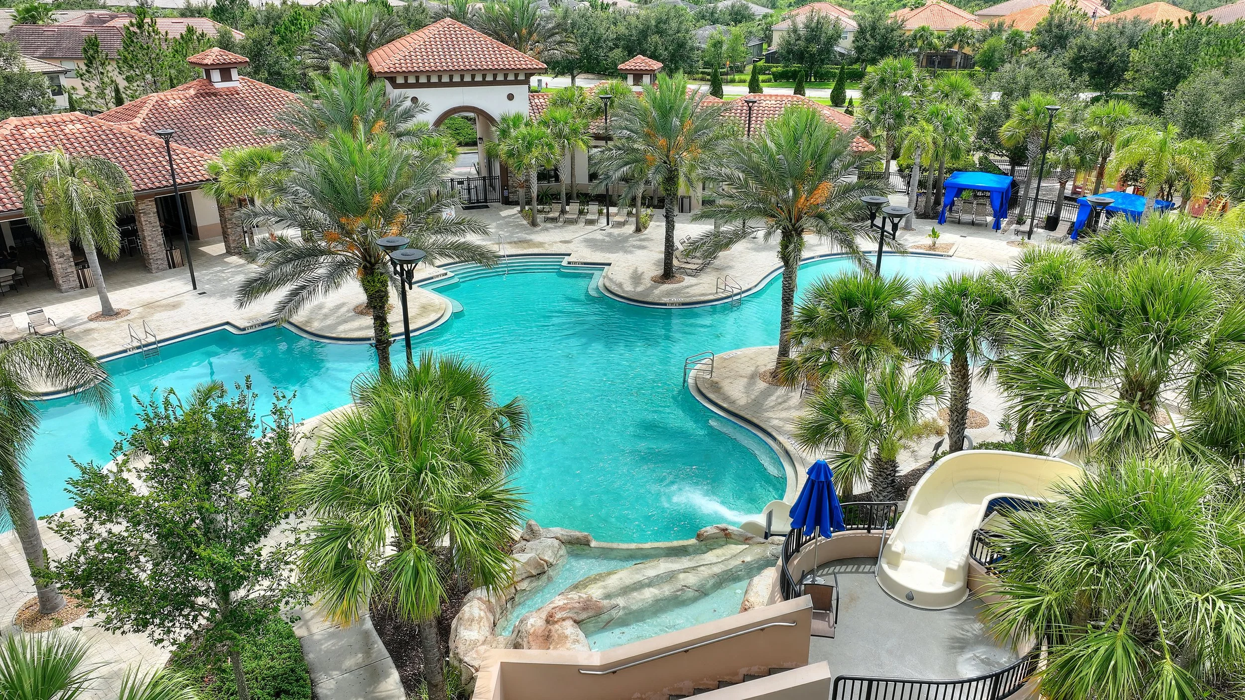 Swimming pool area with a water slide, palm trees, lounge chairs, and cabanas in a resort or community setting.