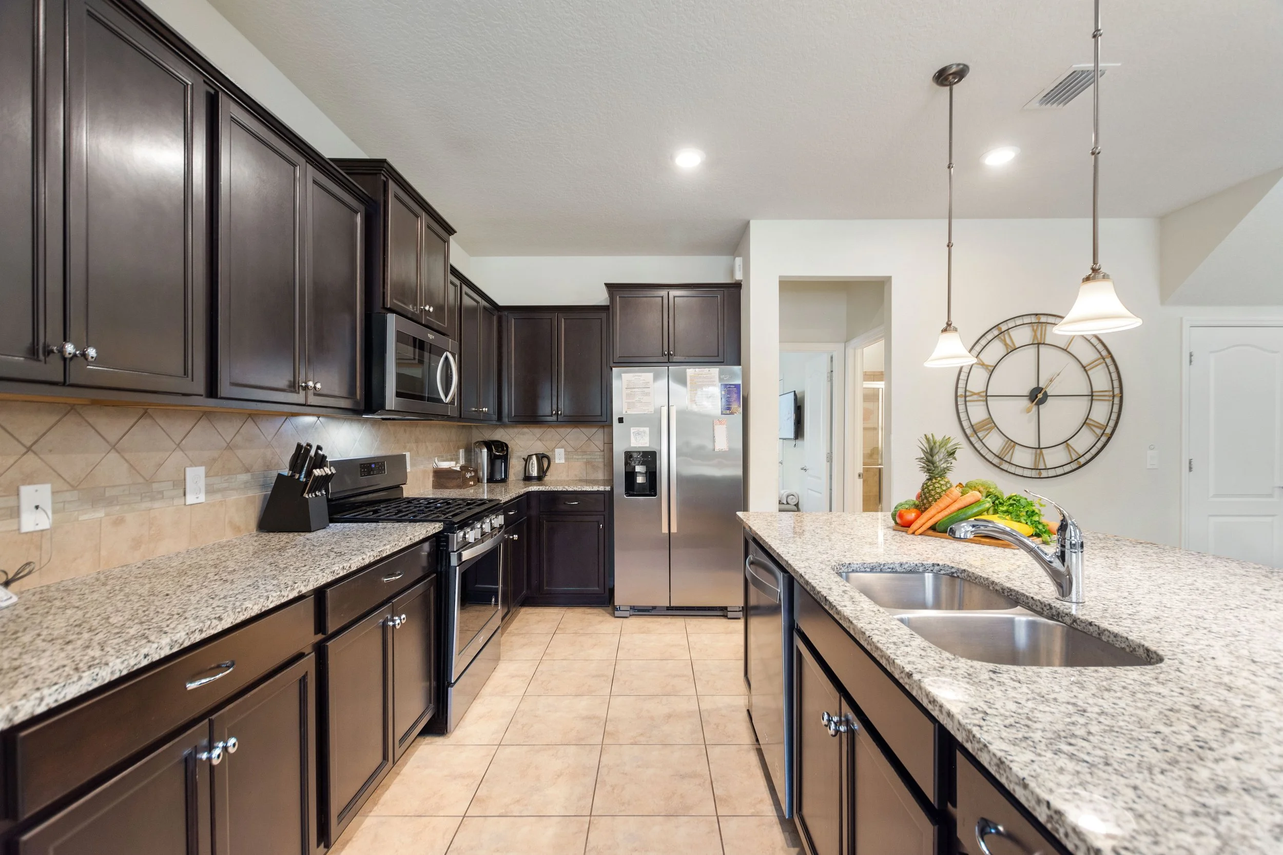 Modern kitchen with dark wood cabinets, granite countertops, stainless steel appliances including a refrigerator, stove, microwave, and coffee maker. There is a double sink on an island with fresh fruits and vegetables. A large wall clock with Roman 