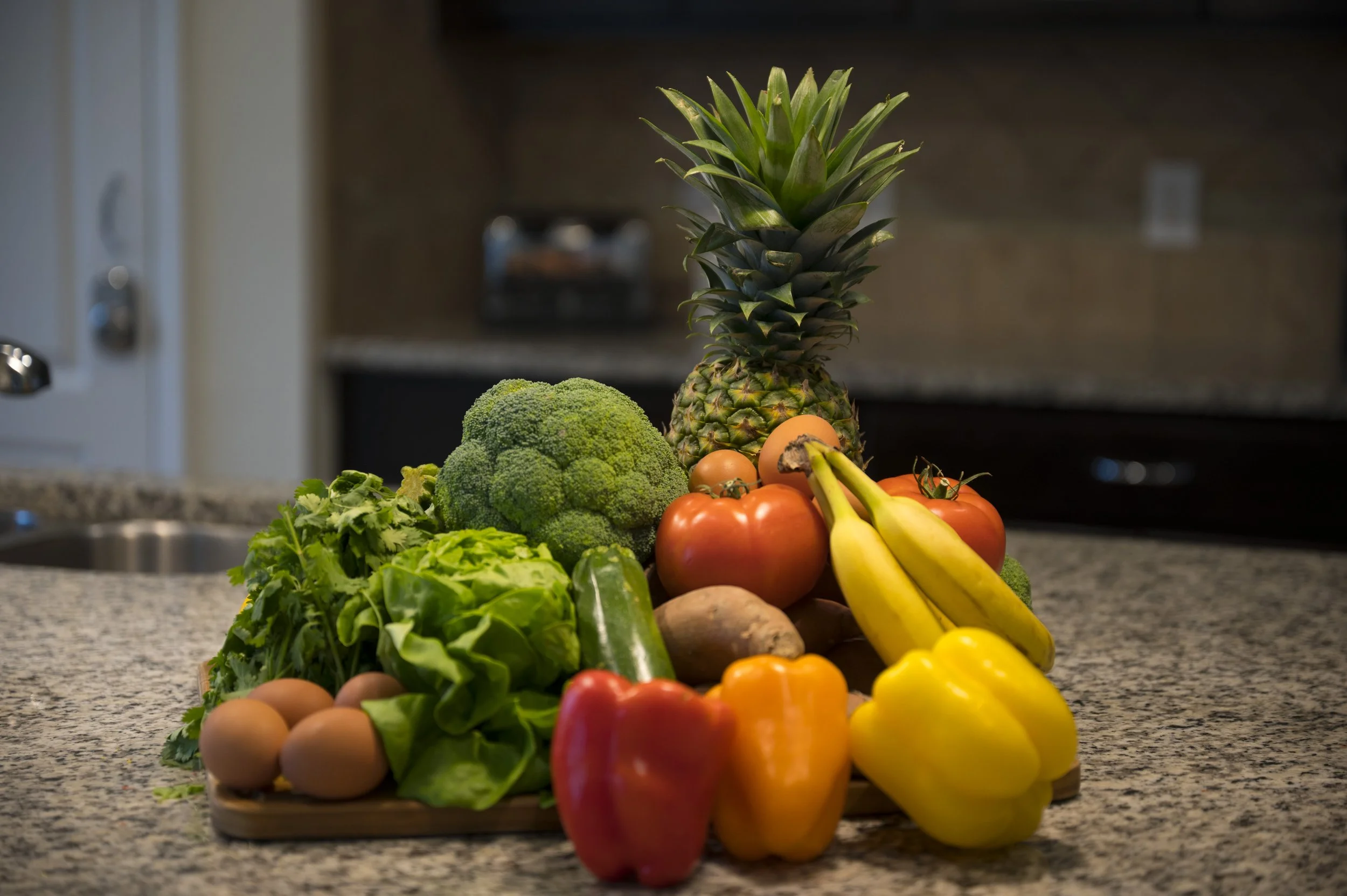 Fresh vegetables and fruits on a granite countertop, including pineapple, broccoli, lettuce, tomatoes, bananas, yellow and red bell peppers, eggs, zucchini, and a potato.