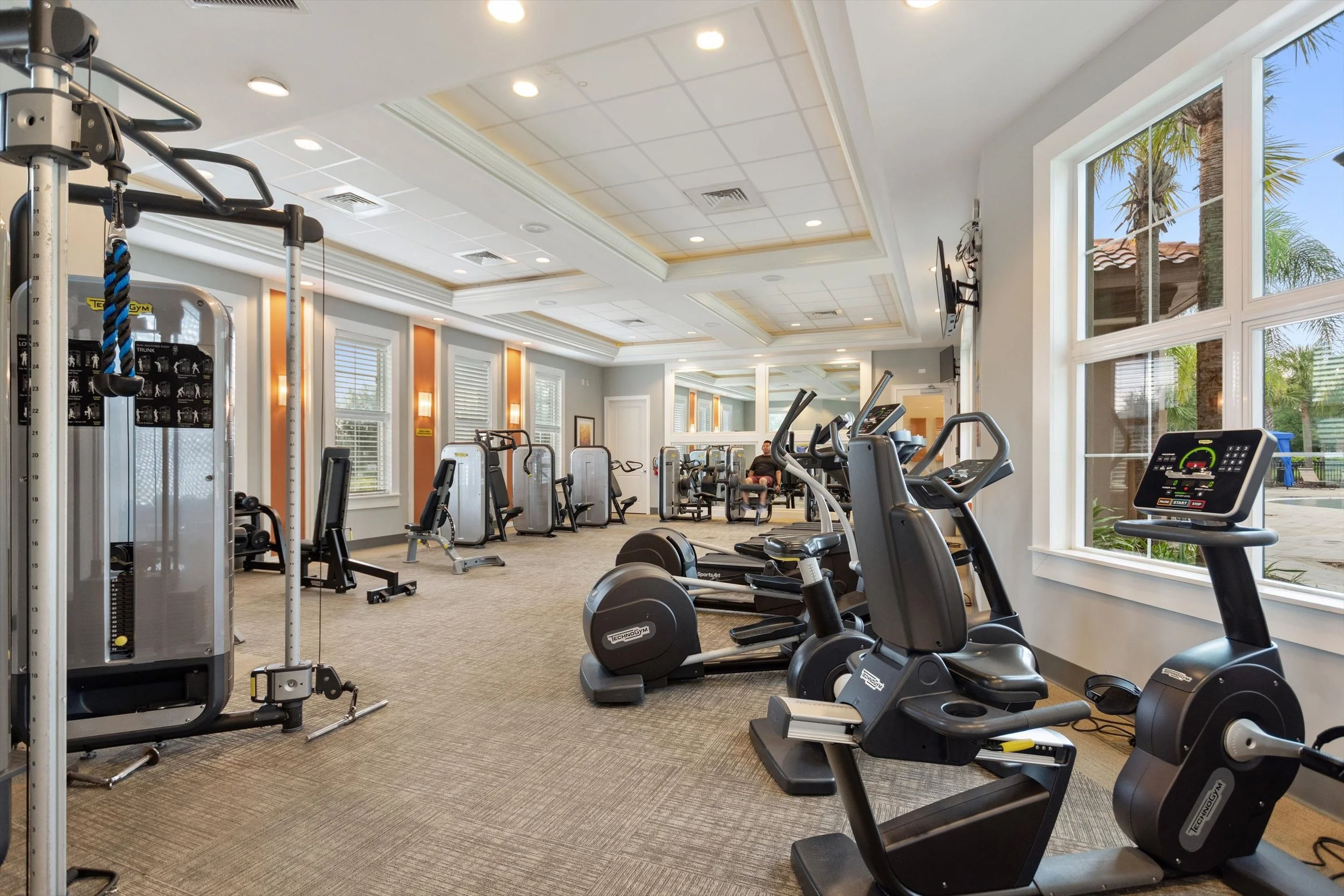 Inside a well-lit gym with various exercise equipment, including stationary bikes, elliptical machines, and weight machines, with large windows showing palm trees outside.