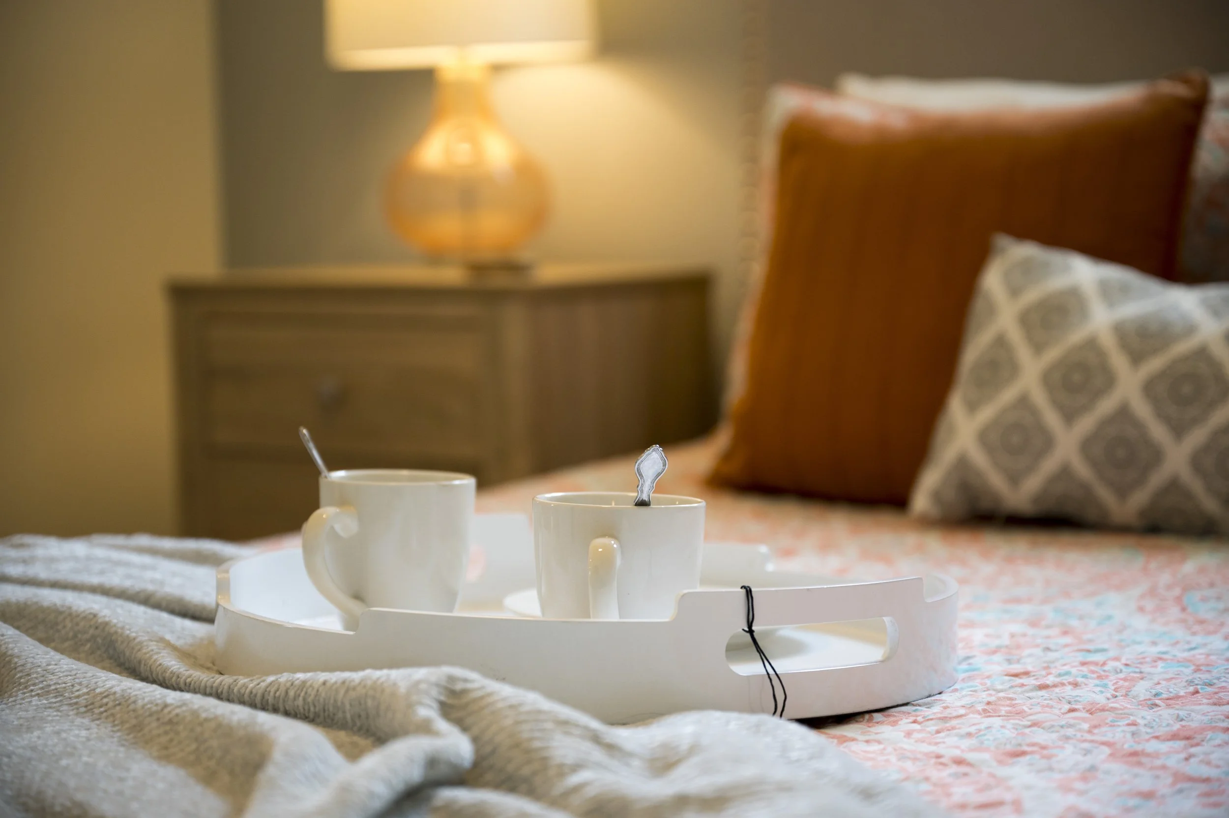 A bedroom scene with a tray holding two coffee mugs with spoons, on a bed with a blanket and pillows, a nightstand with a lamp in the background.