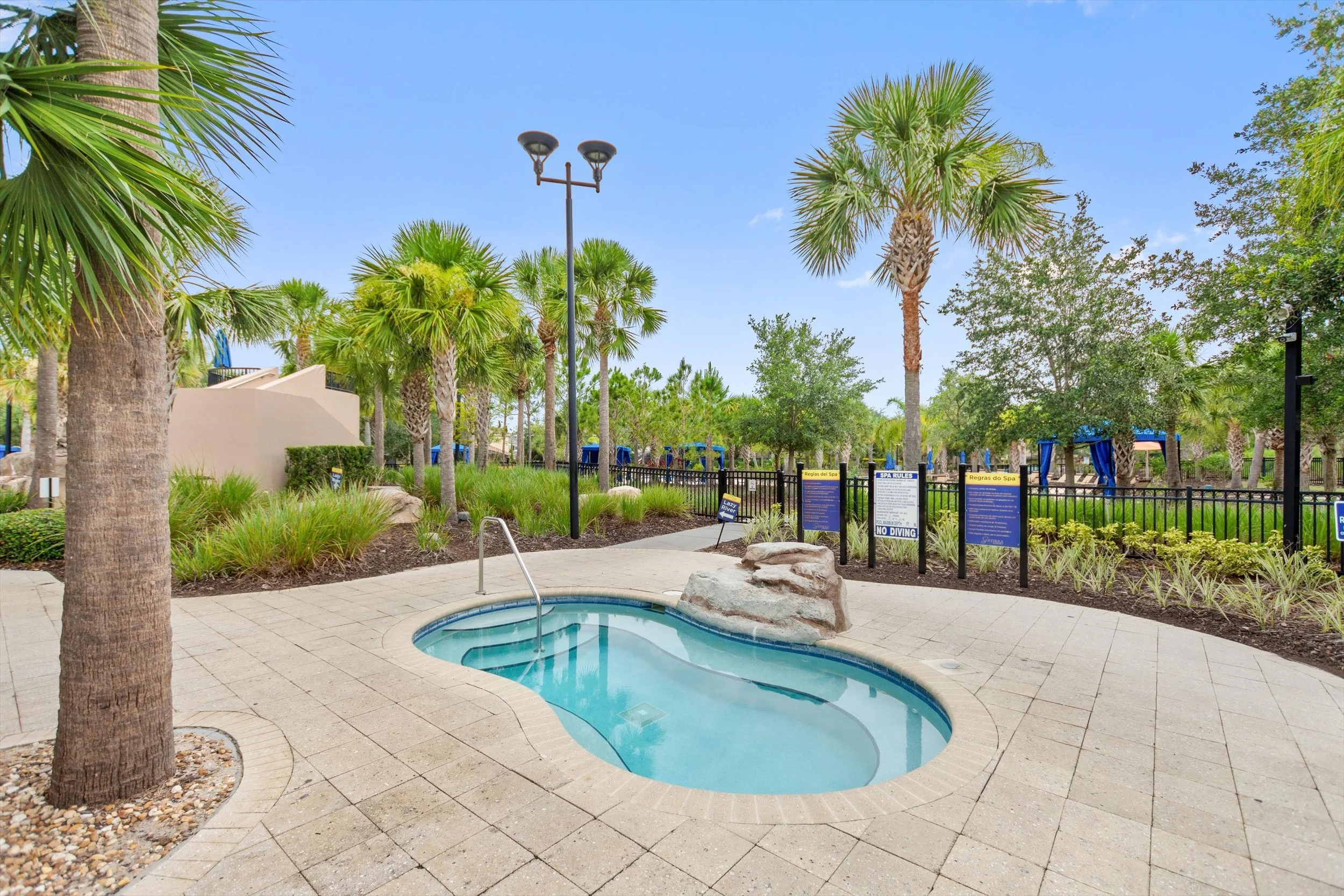 Small pool with a rock at the edge surrounded by tropical palm trees and greenery, with signs and a fence in the background.