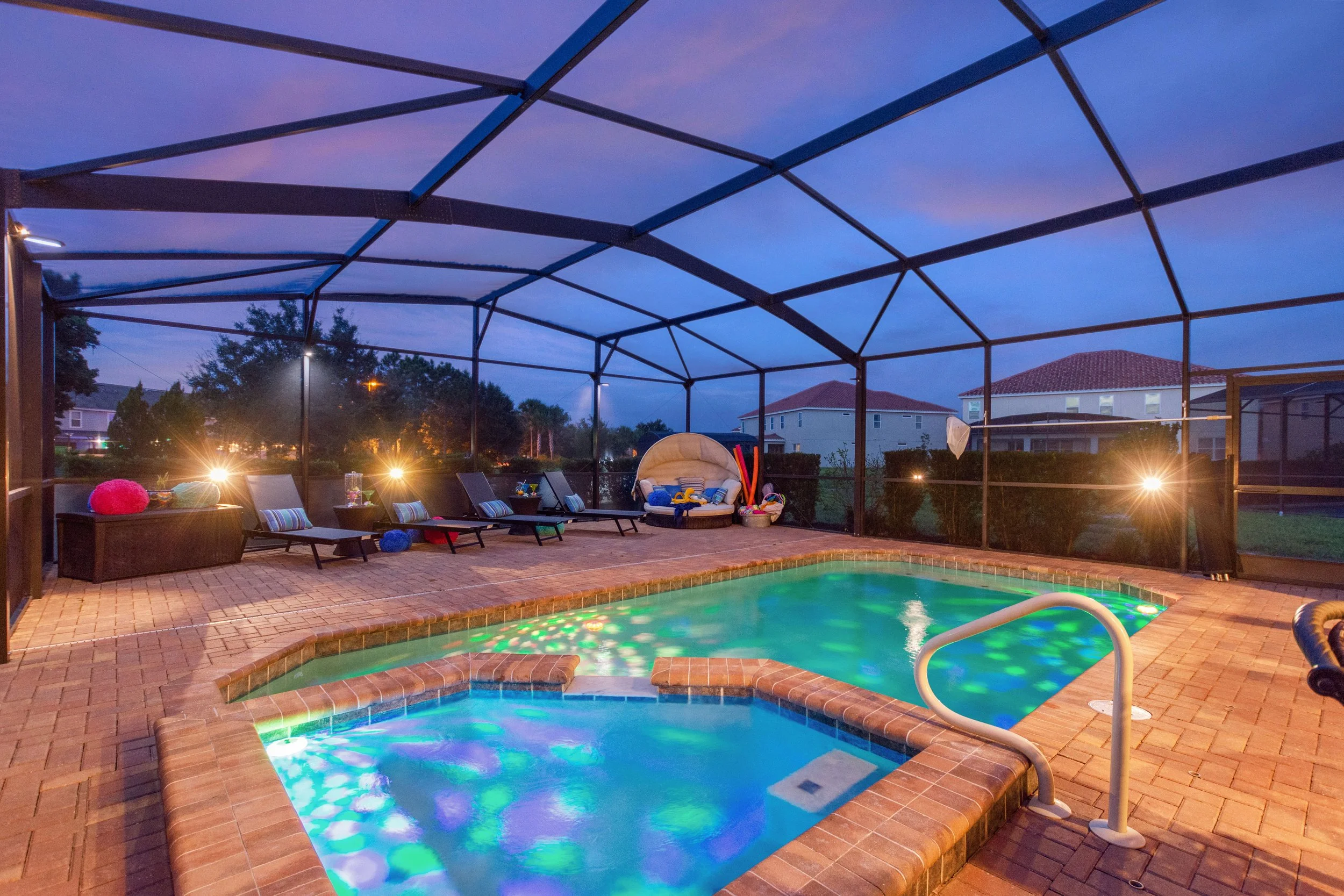 Enclosed backyard with two illuminated swimming pools, outdoor lounge chairs with striped pillows, a round daybed with cushions, and various colorful pool toys. The enclosure has a domed metal frame with mesh screening, and neighboring houses are vis