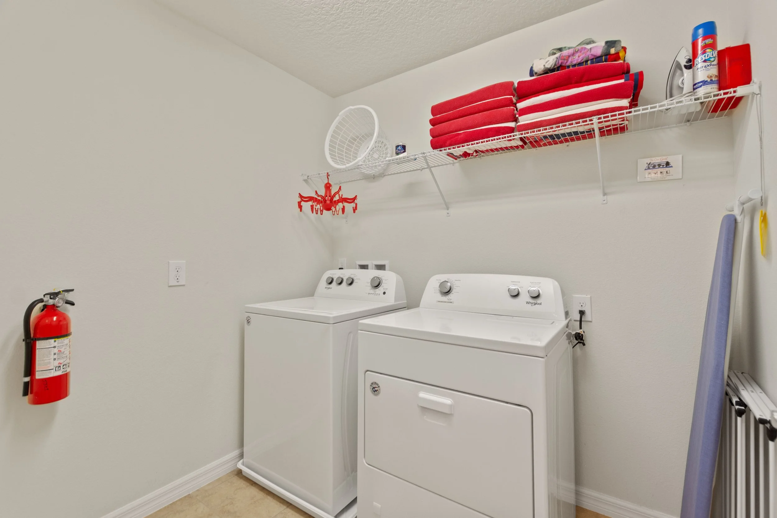 A laundry room with a white washer and dryer, red towels on a wire shelf, a fire extinguisher on the wall, and an ironing board in the corner.