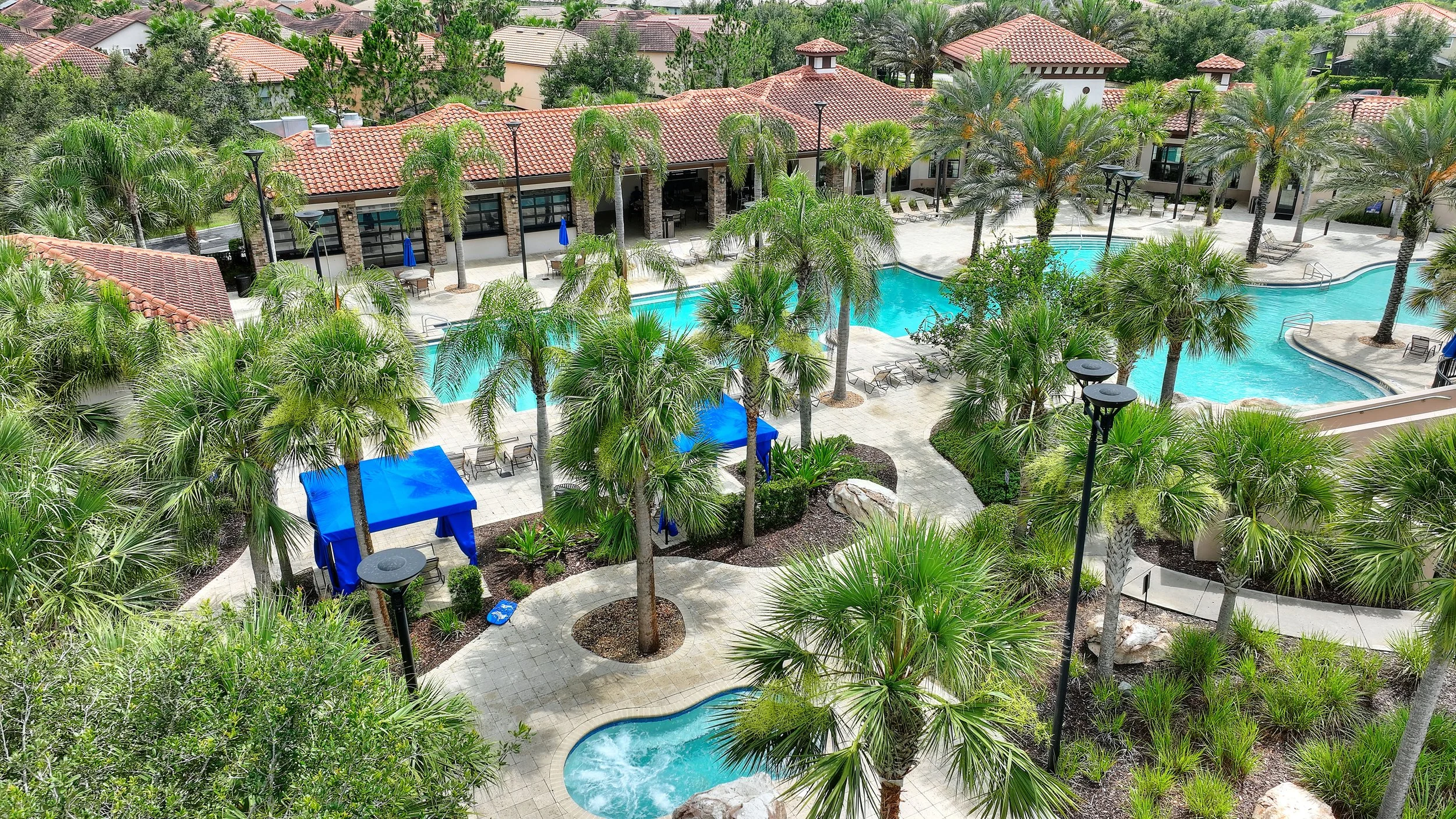 Aerial view of a resort-style swimming pool area with palm trees, lounge chairs, shaded cabanas, and a building with a red tile roof surrounded by greenery.