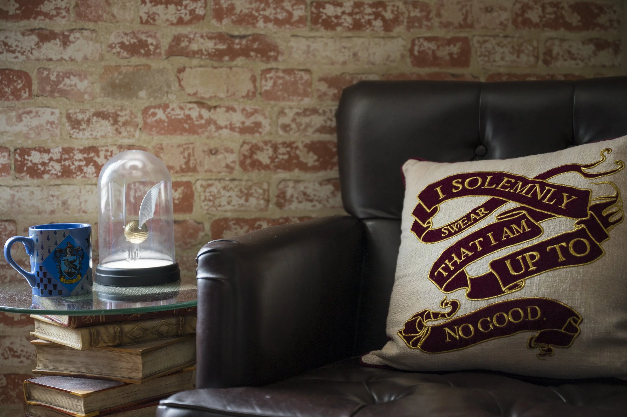 A black leather sofa with a beige pillow embroidered with a quote in maroon and gold. Next to the sofa is a glass-top side table holding a stack of books, a blue mug with cartoon characters, and a decorative glass dome with an object inside, set agai
