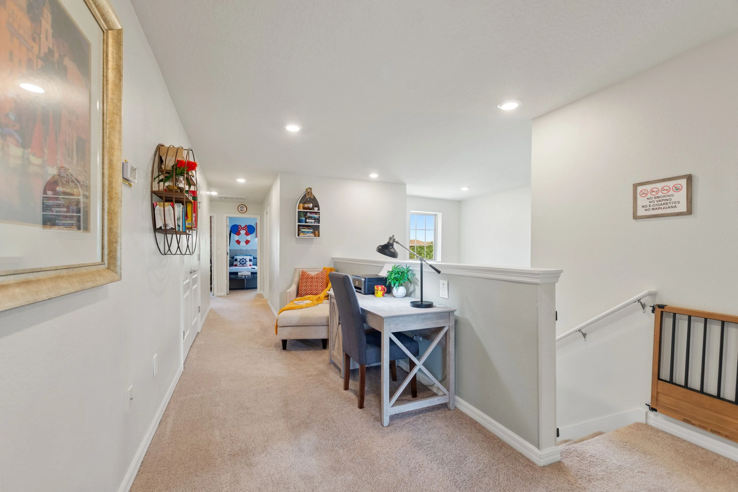 Upscale view of a cozy, well-lit second-floor hallway with beige carpet, white walls, a small sitting area with a chair and yellow throw blanket, a desk with a black lamp, and decorative wall shelves, leading to a bedroom with nautical-themed decor.