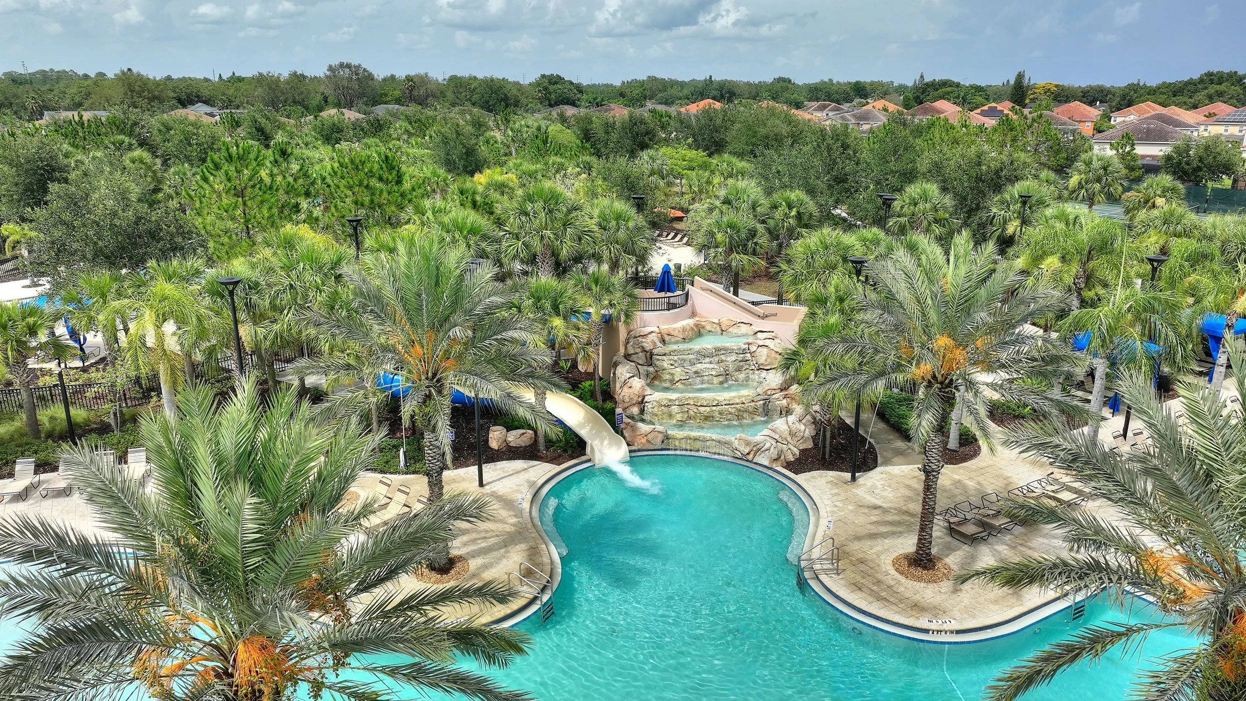 A tropical-themed water park pool area with palm trees, a water slide, a waterfall feature, lounge chairs, and a fenced-off relaxation zone.