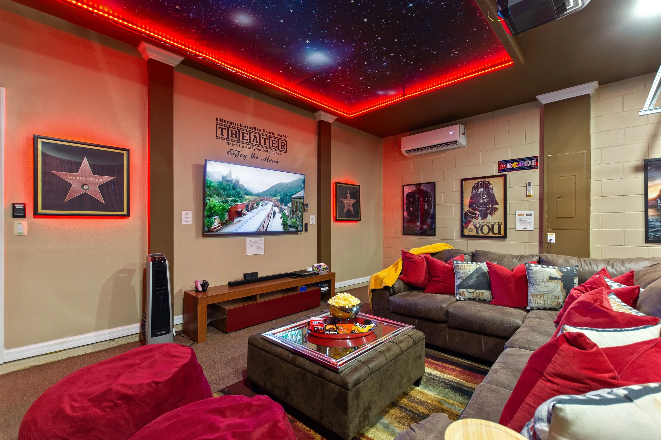 A cozy living room with a starry sky ceiling, red LED lighting, a large L-shaped brown sofa with red and patterned pillows, a glass-topped ottoman with snacks, and a wall-mounted TV showing a scenic view. The walls have movie-themed posters and a sma