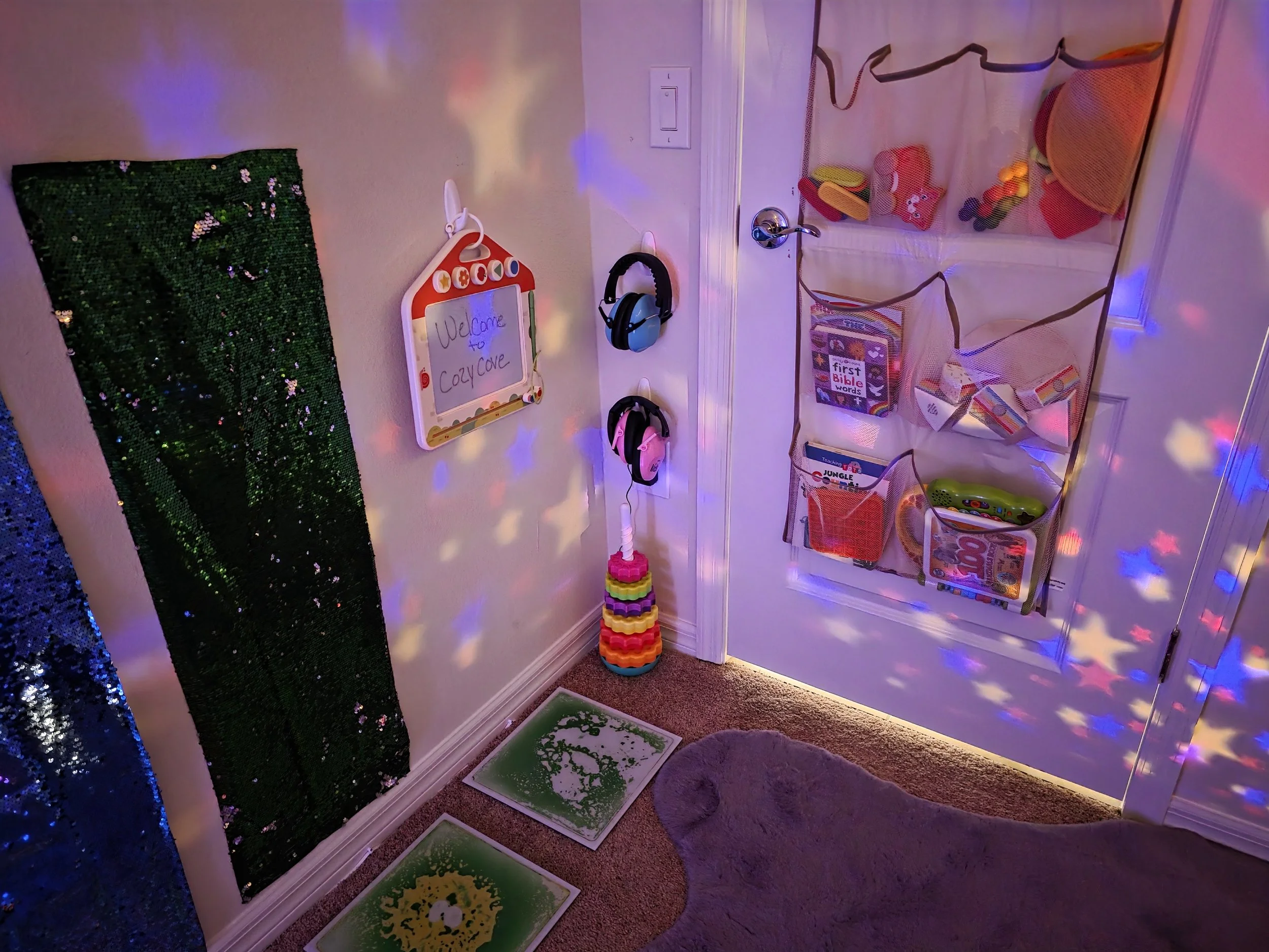 Jordan's Room at Anchor Cove Villa -- entry area with noise-cancelling headphones, a sequin sensory panel, a welcome sign, colorful stacking rings, sensory activity mats, a soft animal rug, and a door organizer filled with books and soft toys.
