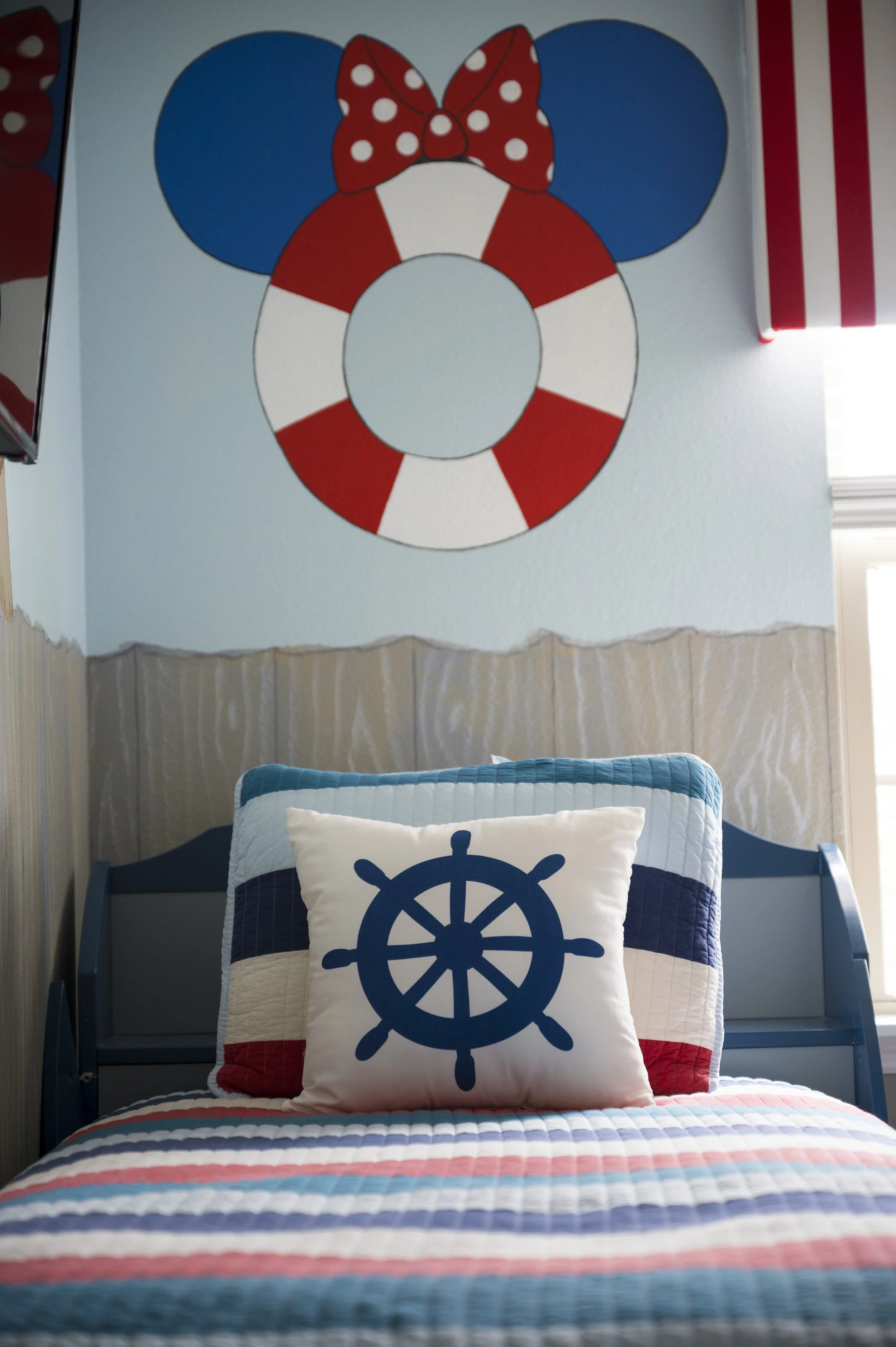 A bedroom with nautical and beach-themed decor, featuring a bed with a quilted striped bedspread and pillow with a ship wheel design, and a wall decoration of a lifebuoy with a red bow and Mickey Mouse ears above the bed.