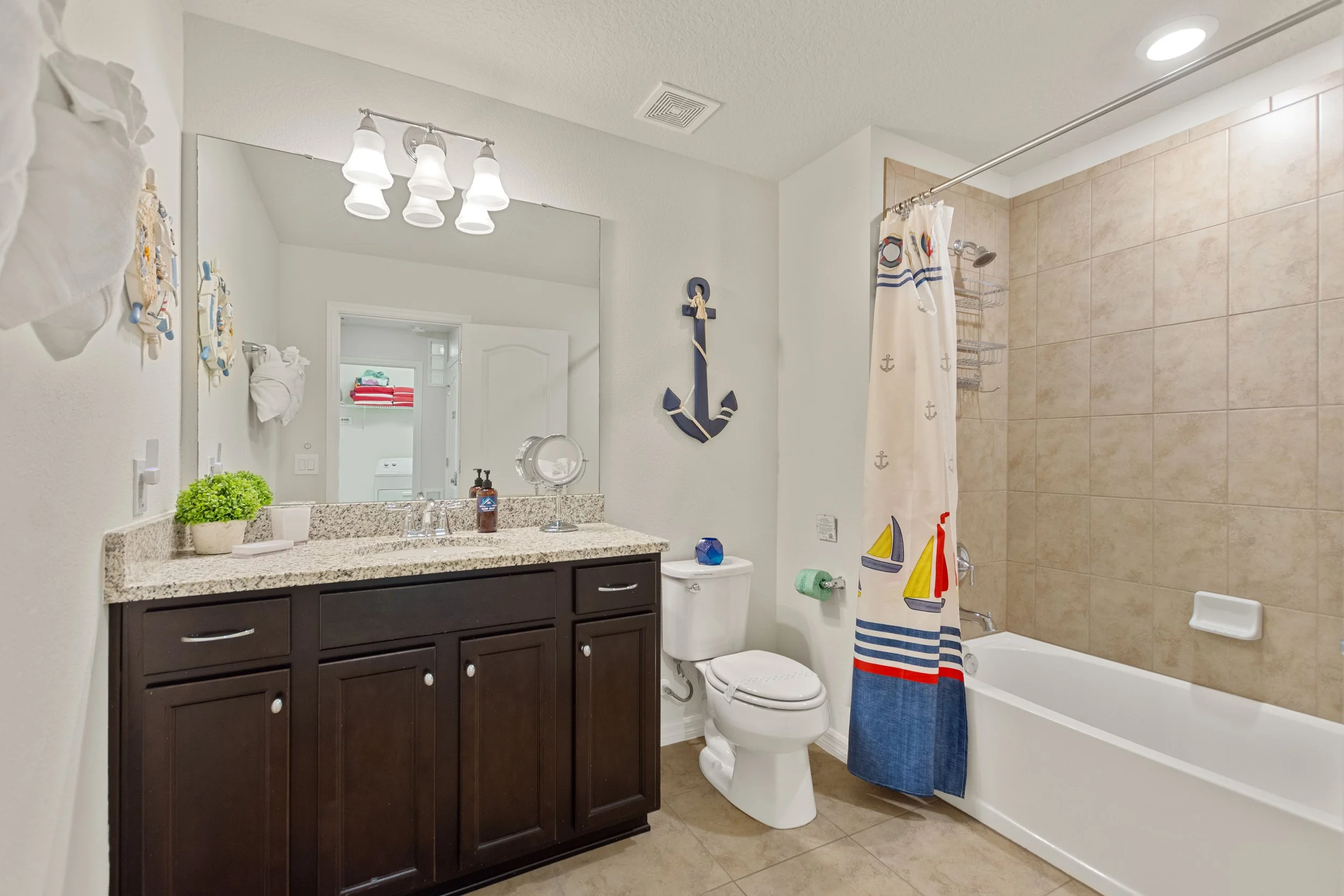 Bathroom with a dark wood vanity, granite countertop, wall mirror, light fixture, toilet, bathtub with nautical-themed shower curtain, anchor wall decor, and laundry area visible through an open door.