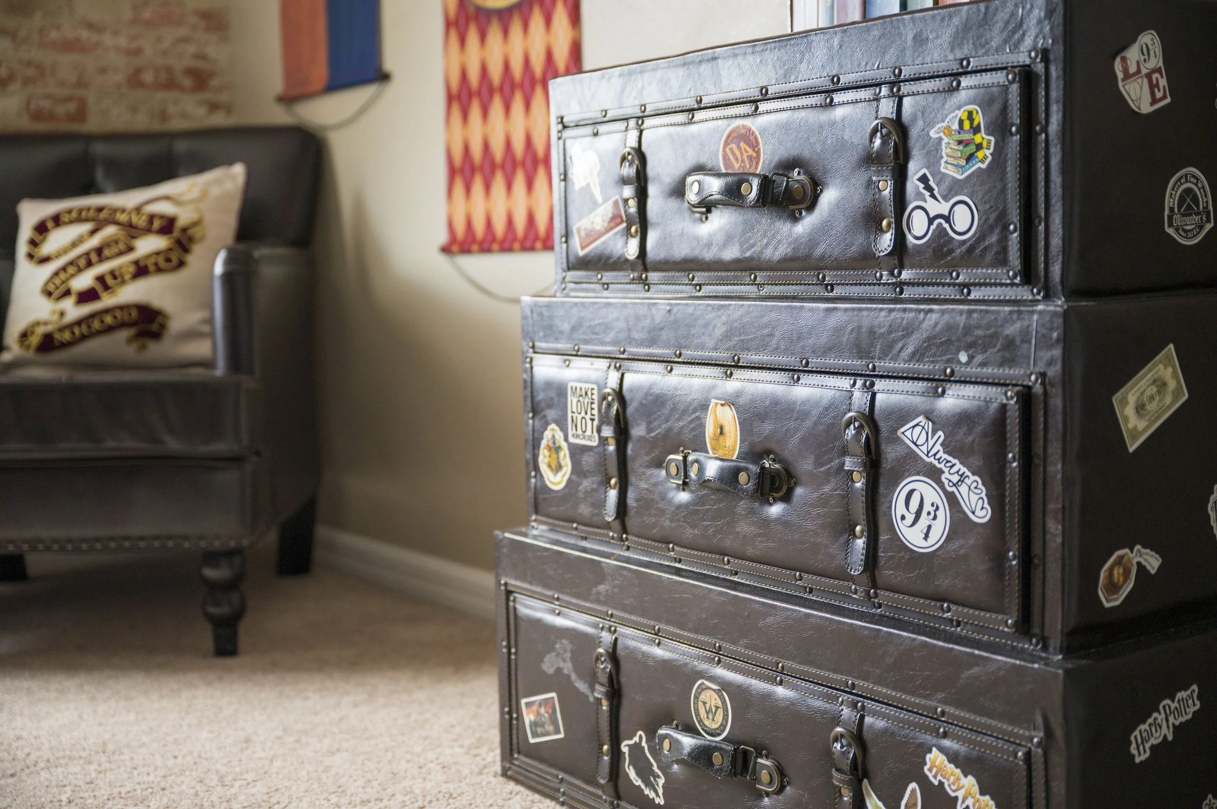Stacked vintage leather suitcases decorated with Harry Potter and other themed stickers, on beige carpet next to a black leather armchair with a Hogwarts-themed pillow, in a cozy room with wall art.
