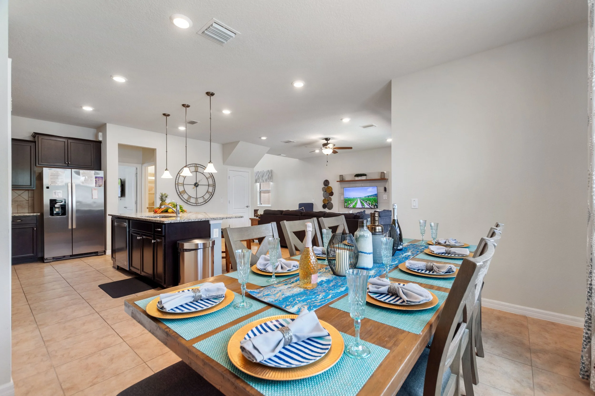 Open-concept kitchen and dining area with a wooden dining table set for six, featuring striped blue and white plates, napkins, and glassware. The kitchen has dark cabinets, a granite island, and stainless steel appliances, with pendant lights hanging