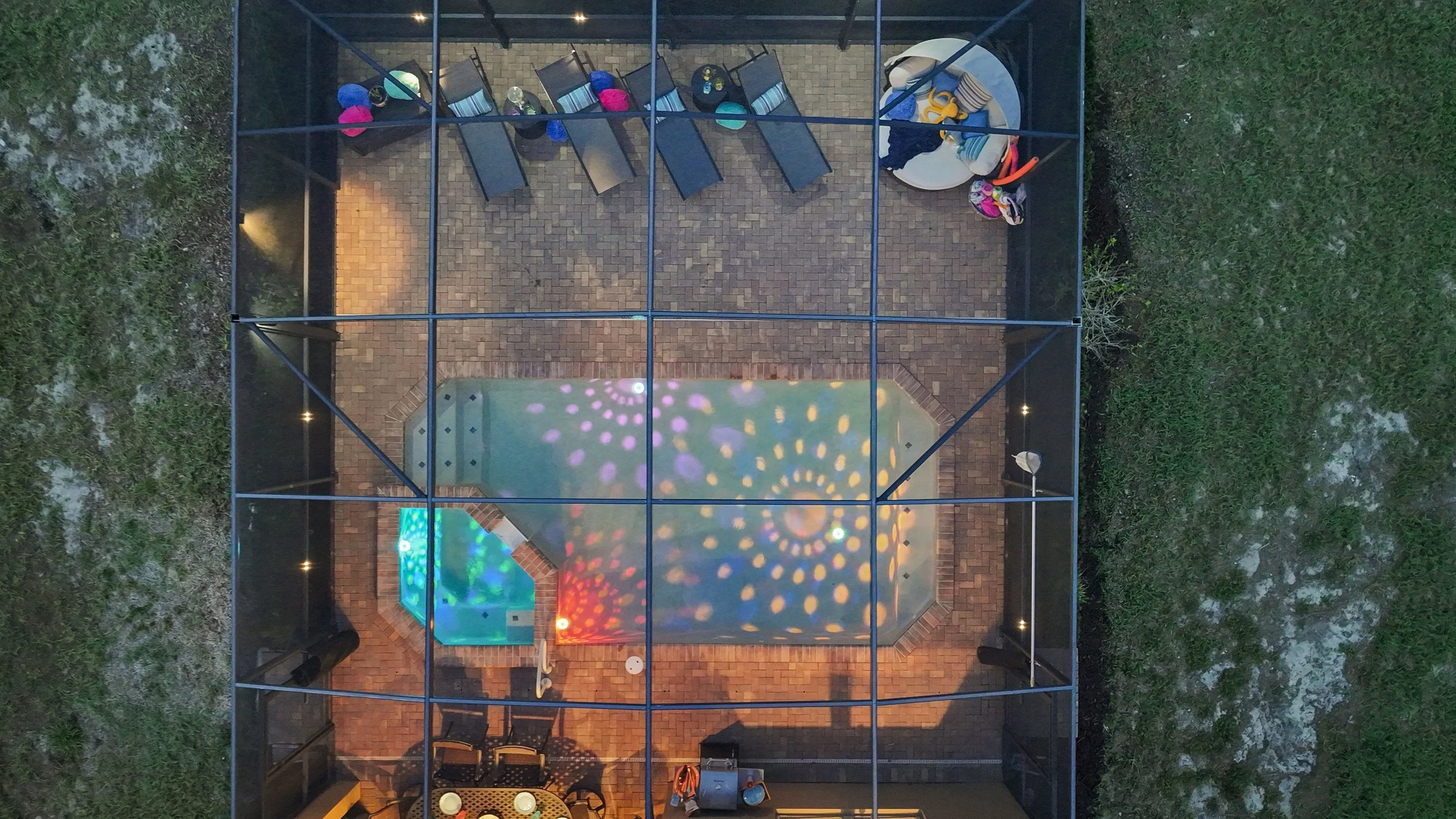 An aerial view of a screened-in patio with a swimming pool, hot tub, poolside chairs, and pool toys. Disco ball light reflections are visible on the pool surface, and outdoor lighting illuminates the patio.