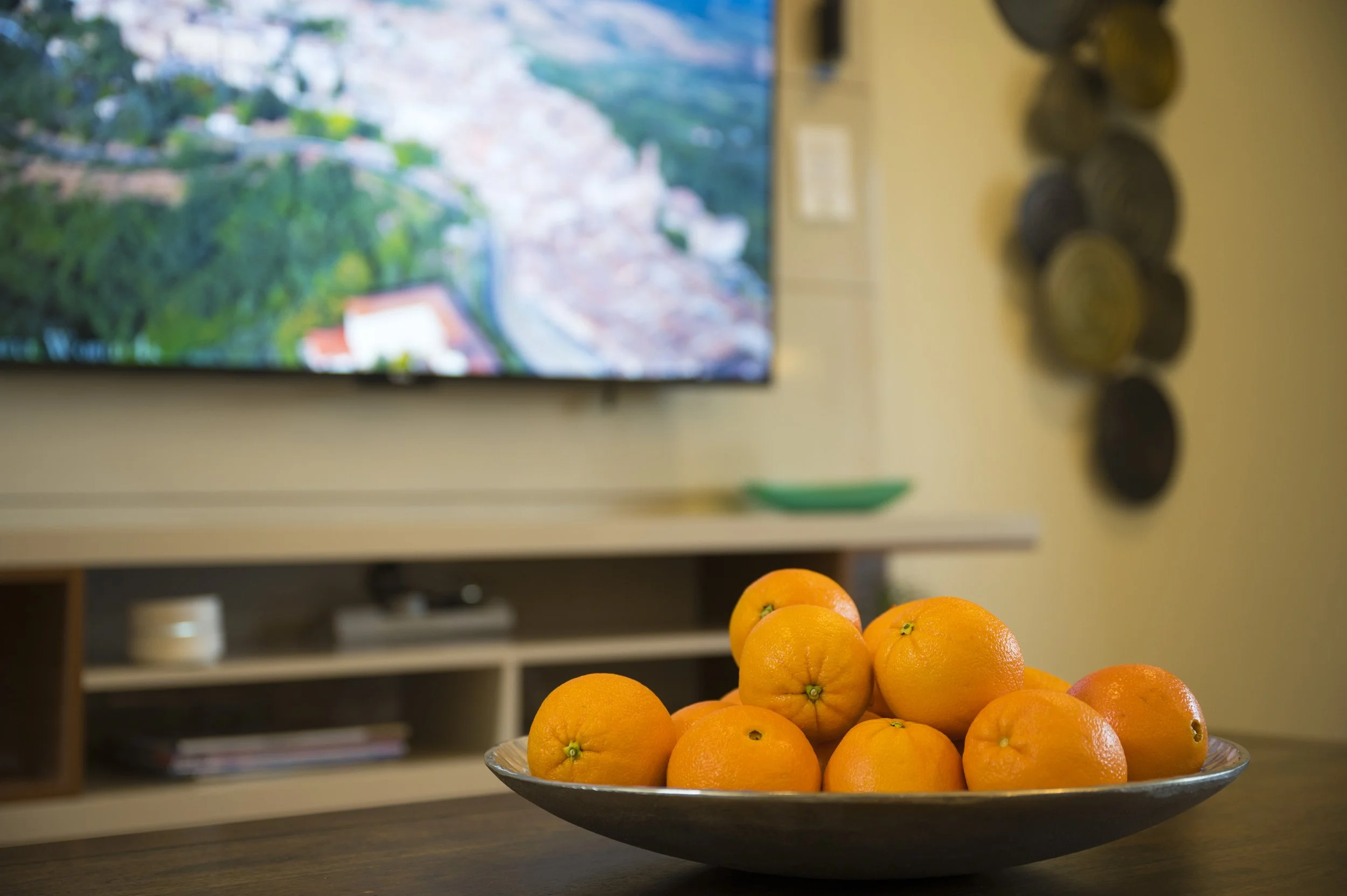 A bowl of oranges on a table in front of a blurred television in a living room.